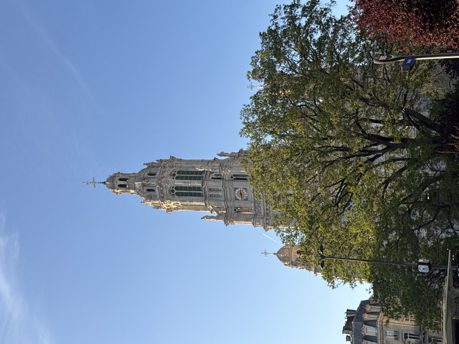 The image captures a daytime scene dominated by the ornate bell tower of Église Saint-Augustin, located at the intersection of Boulevard Malesherbes and Boulevard Haussmann in the 8th arrondissement of Paris, France.
The central subject is the tall, light-colored stone tower, characterized by multiple architectural tiers, classical columns, arched windows with dark latticework, and various sculptural elements. A circular clock face with black Roman numerals is visible on one side of the tower. The tower culminates in an elaborate spire topped with a cross. To the left of the main tower, a smaller, domed section of the church, also crowned with a cross, is partially visible.
The foreground and mid-ground feature dense foliage from several trees. A large tree with bright green leaves partially obscures the lower sections of the church, while a tree with dark reddish-purple leaves is visible on the far right. A black double-headed street lamp is positioned in the lower-left-center. To the left, a multi-story, light-colored urban building with dark roofs and dormer windows, typical of Parisian Haussmannian architecture, is partially visible. On the right, partially obscured by the red-leaved tree, is a vertical black pole with a blue circular sign indicating a bus stop.
The sky is a clear, bright blue, with a faint white contrail visible high above and to the left of the main tower. The scene is illuminated by strong, direct sunlight, casting minimal shadows. No people or discernible actions are present.