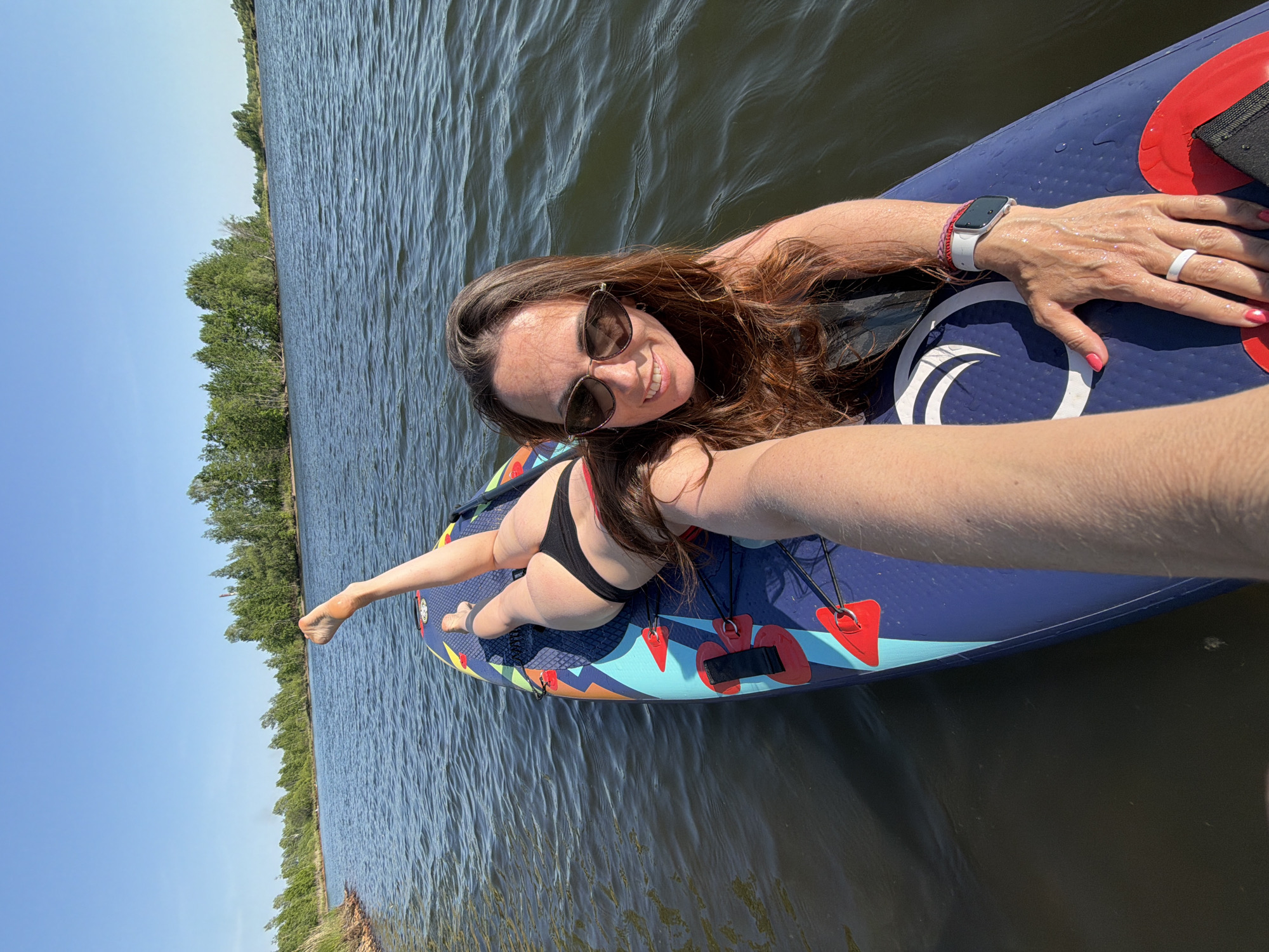 A woman is lying on a paddleboard in the water. She is wearing sunglasses and a black bikini. She has a watch on her left wrist and a ring on her left hand. The paddleboard is blue and has red and yellow designs. The water is dark and there are trees visible in the background. The location is Razdolnoye, Russia.