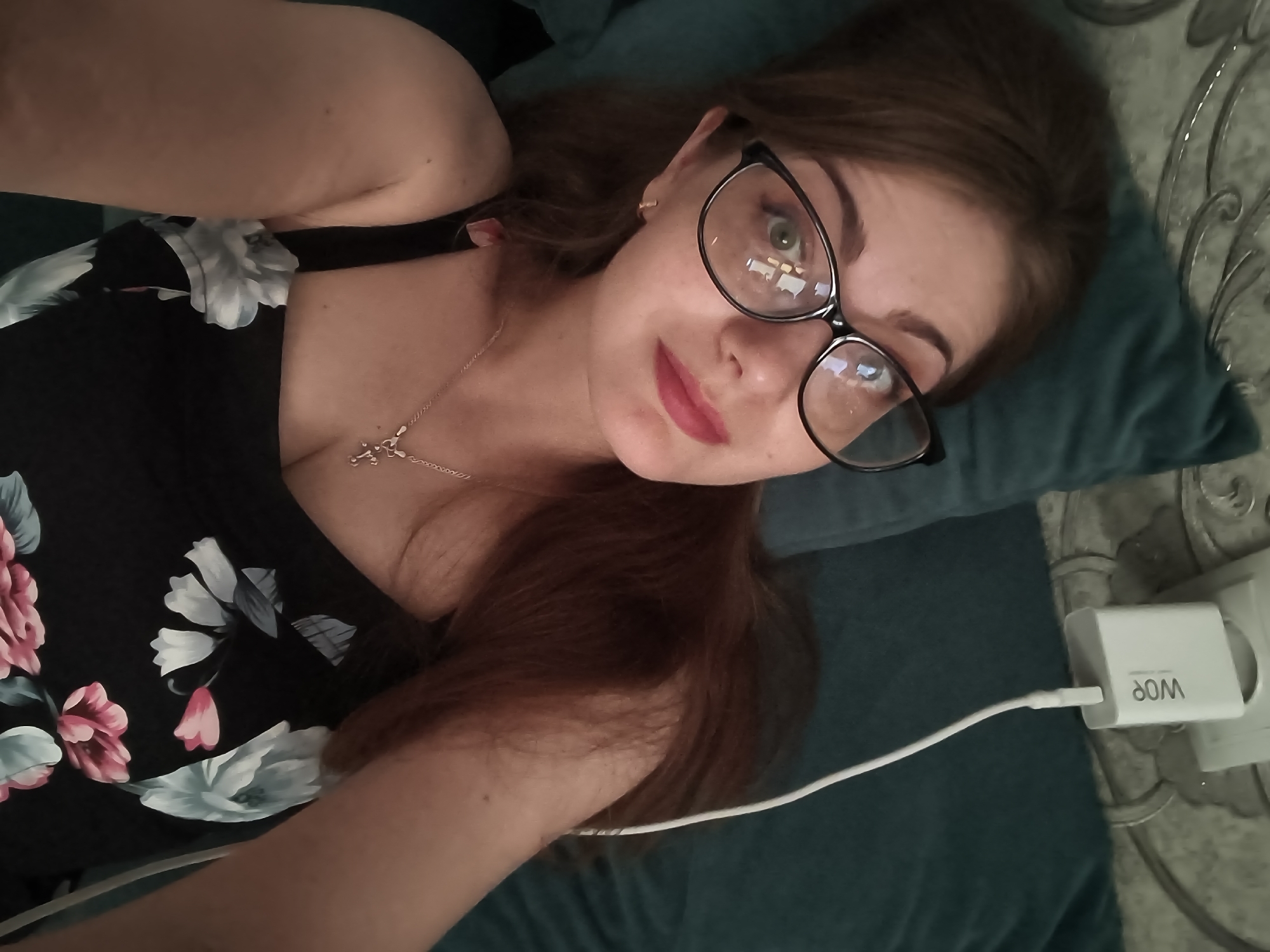 A woman is lying on a pillow, looking at the camera. She has long brown hair and is wearing glasses and a black dress with a floral pattern. Her right arm is bent at the elbow and her hand is near her face. A white phone charger is plugged into an outlet in the lower right corner of the image. The woman is in a room with a patterned wall on the right side of the frame.