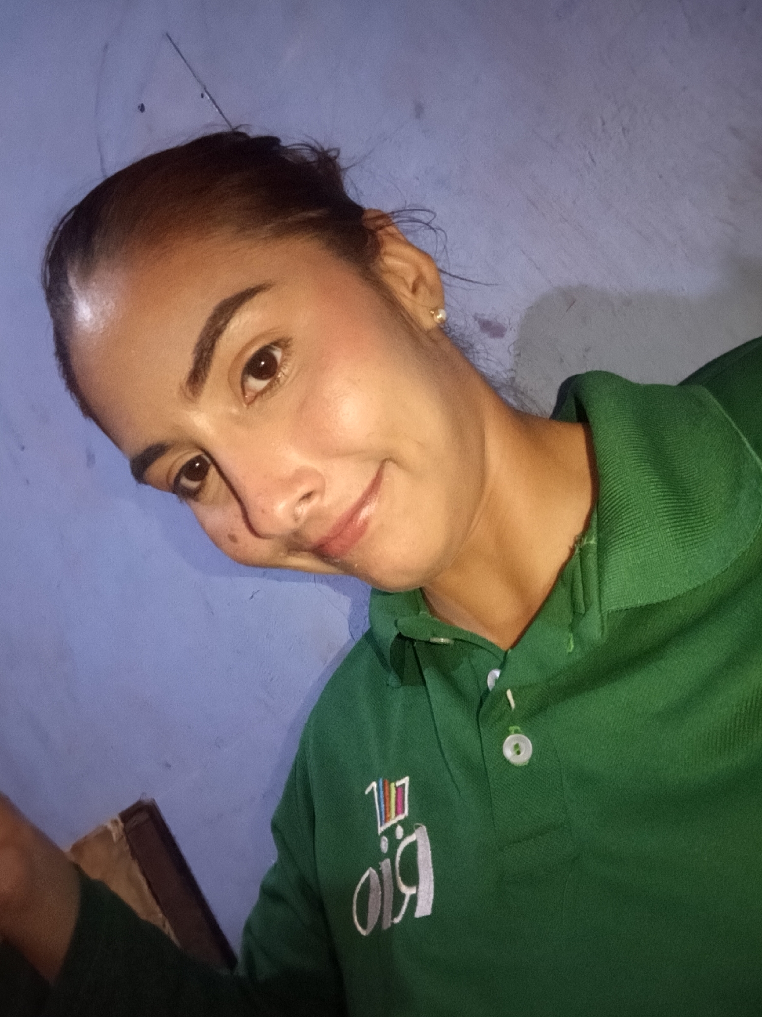 A woman with tan skin and brown hair pulled back in a bun poses indoors against a mottled blue wall. She wears a green collared shirt with a white button and a partially obscured white embroidered design on the left breast. She is angled to the left, looking at the camera with a slight smile. She wears a stud earring in her right ear. A piece of furniture, possibly a chair, is visible in the lower left corner. There is no indication of the location being Juan Griego, Venezuela or any specific location.