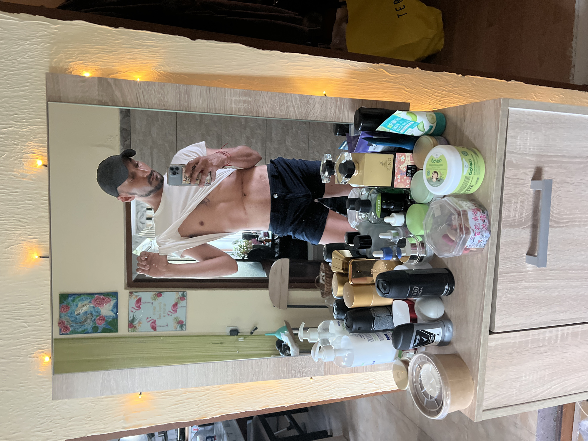 The scene depicts an adult male taking a mirror selfie in a room in Medkovets, Bulgaria. The male, appearing to be in his 30s-40s, is wearing a black baseball cap, dark shorts, and a white t-shirt which he holds up with his left hand to expose his upper abdomen. Several horizontal scars are visible across his mid-upper abdomen. He holds a light blue iPhone in his right hand, pointing it towards the mirror to capture his reflection. A red string bracelet is visible on his left wrist. The dresser in the foreground, made of light wood-grain material with a silver-colored handle, is covered with numerous personal care products including various pump bottles, two deodorant cans (one labeled "Rexona Original," another "R8 ORIGINAL"), several perfume bottles (one gold labeled "ZENO," another dark with "VETIVER"), multiple jars of cream (one green labeled "БОЧКО"), dropper bottles, and other cosmetic items.
Reflected in the mirror, behind the male, are two framed pictures on a light-colored, textured wall: the top picture features pink flowers and fish on a blue background, while the bottom picture displays pink flamingos and tropical leaves on a light blue background, with some text partially visible. To the left in the reflection, part of a dining area with a table and chairs is visible, along with a green curtain and a white object resembling a dolphin toy on a shelf. To the right in the reflection, a dark brown curtain, a partially visible yellow bag (showing "TED B..."), and a wooden stool or chair are discernible. Small, warm-toned string lights are attached to the textured frame of the mirror or the wall above it. The floor visible in the reflection is light-colored.