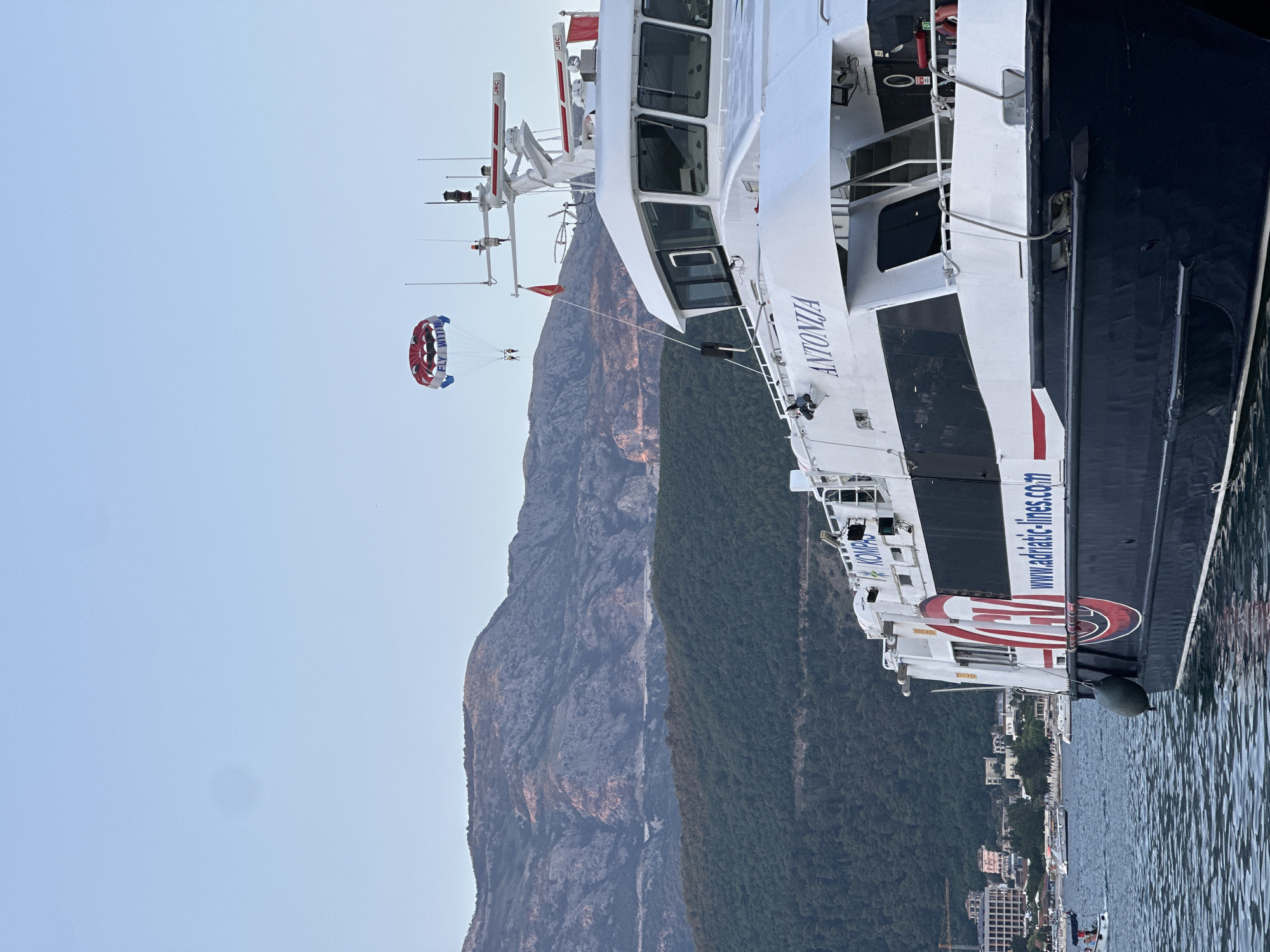 The image depicts a parasailer being towed in the sky above a ferryboat named "ANTONIJA" docked in the sea. The ferryboat has a dark blue hull and a white superstructure with the text "www.adriatic-lines.com" visible on its side. In the background, a city with buildings and lush green hills rise up to meet rugged, rocky mountains under a clear blue sky. The scene appears to be set in Budva, Montenegro, a coastal town known for its scenic beauty and recreational activities. The parasailer, a colorful canopy with two people suspended below, is positioned in the upper portion of the image, indicating it is engaged in a recreational activity. The ferryboat dominates the foreground, suggesting it is a prominent feature of the harbor. The presence of other boats in the distance and the calm water indicate a peaceful setting.