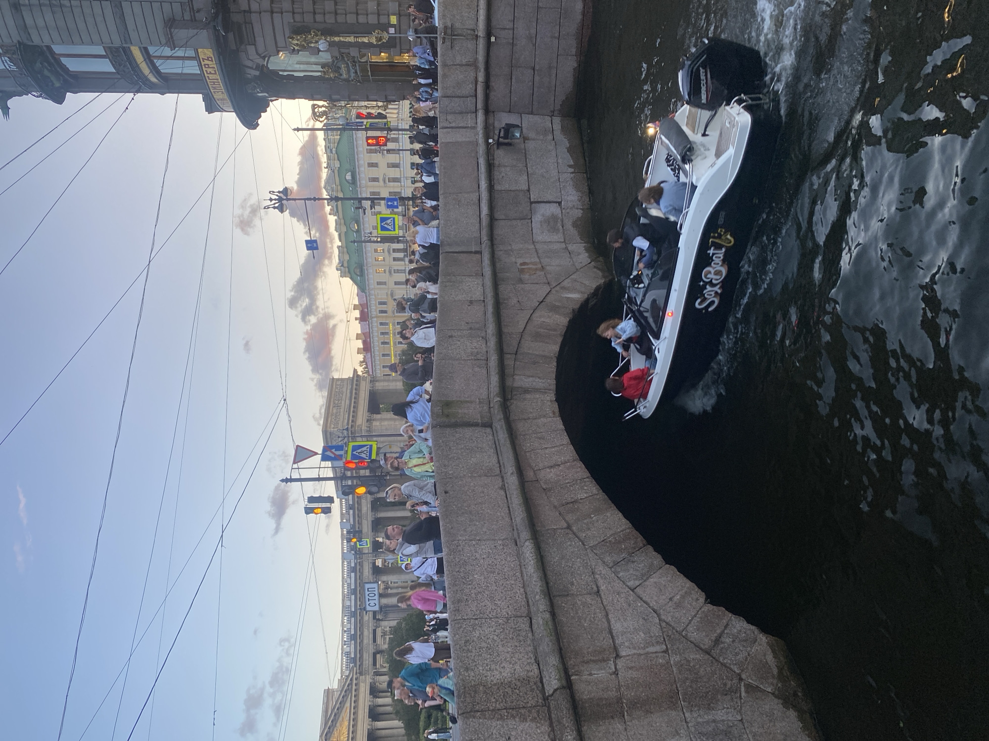The image shows a view from a canal in Saint Petersburg, Russia. In the foreground, a motorboat with "Sax Boat" written on its side is moving through the dark water, leaving a wake. Several people are visible on the boat, including individuals wearing red, blue, and grey clothing. The boat is passing under a stone bridge, which is a typical architectural feature of Saint Petersburg's canals.
On the bridge above, a crowd of people is gathered, looking out at the canal or interacting with each other. They are dressed in casual attire. Behind the bridge, a street scene is visible with buildings, traffic lights, and street signs, including one with the Cyrillic word "СТОП" (STOP). The architecture of the buildings suggests a historical city center, consistent with Saint Petersburg. The sky is overcast with a hint of sunset colors. The overall atmosphere appears to be a pleasant evening in a vibrant urban environment.