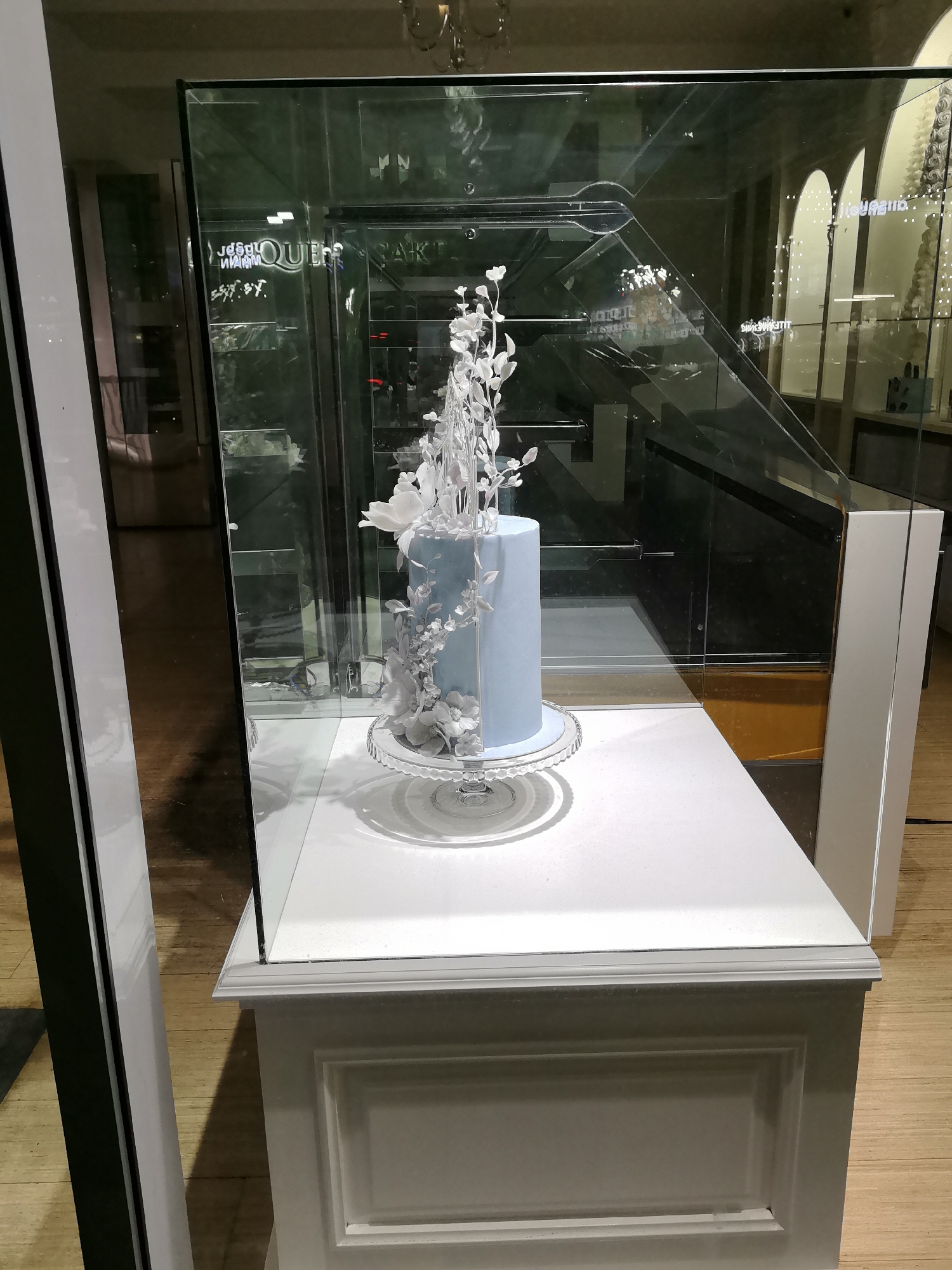 A light blue, cylindrical cake adorned with white floral decorations stands on a clear glass cake stand. The cake is displayed inside a large, glass-enclosed cabinet. The cabinet is positioned on a white pedestal. Reflections in the glass suggest the cake is in a shop or gallery. The background is obscured by reflections and dark surfaces. The floor appears to be made of wood. The overall scene is clean and well-lit, focusing attention on the cake. The location is a shop in Yerevan, Armenia.
