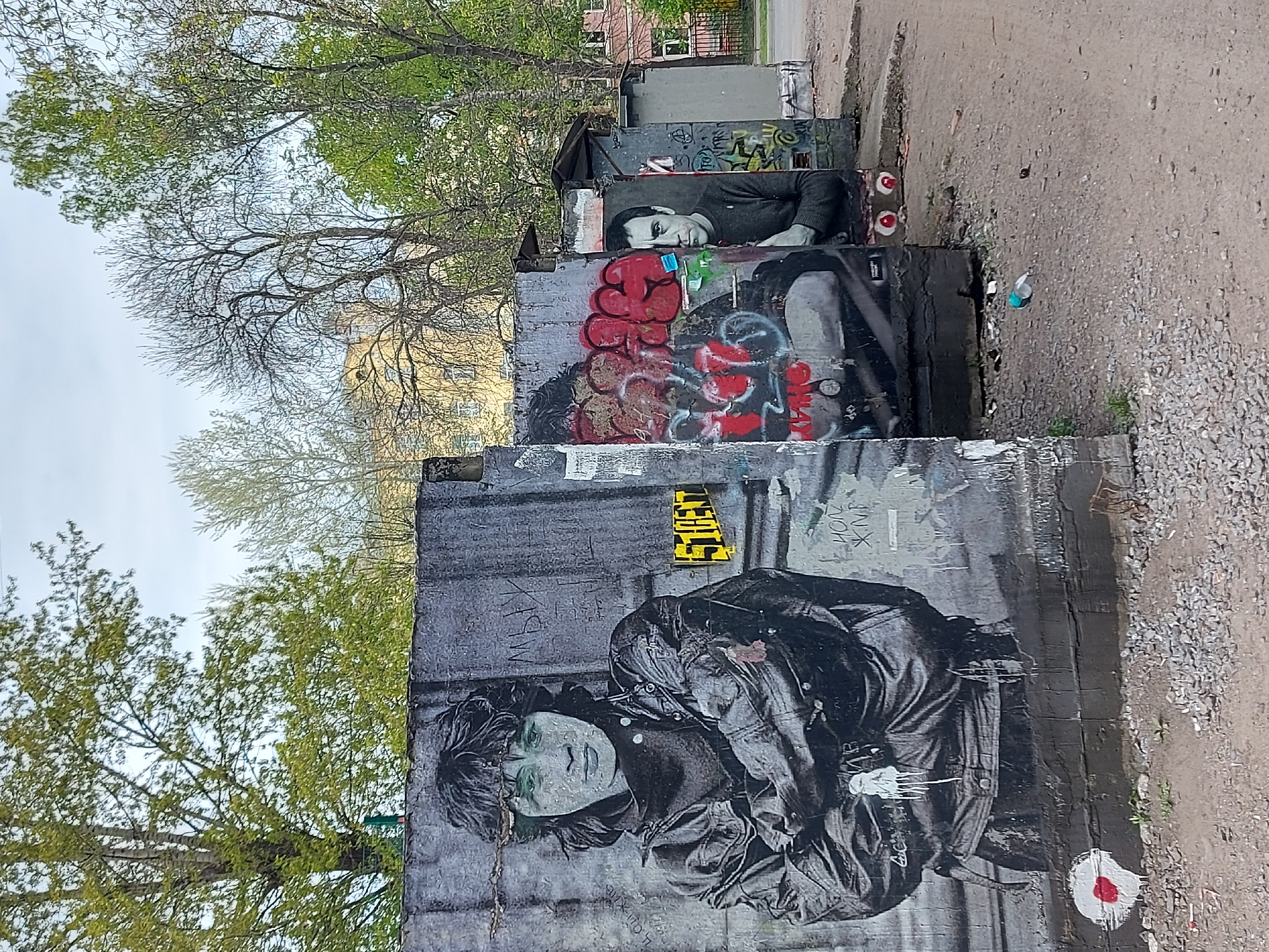 The image shows a series of concrete utility boxes covered in street art, including painted portraits and graffiti. The location is likely Saint Petersburg, Russia, evidenced by Russian language script visible on one of the boxes and the artistic style associated with the city's street art scene.
The lowest box features a large grayscale portrait of a person with dark hair and wearing a leather jacket. There is additional graffiti and paint splatters overlaid on the image. Above it, another box displays a portrait heavily obscured by red and black graffiti. The highest box is decorated with a grayscale portrait of a person with dark hair. Graffiti is present across these boxes.
The boxes are situated near a pedestrian area covered in gravel and dirt. A street runs parallel to the boxes, and a few other buildings are visible. Trees with budding leaves are visible in the background. The sky is overcast.