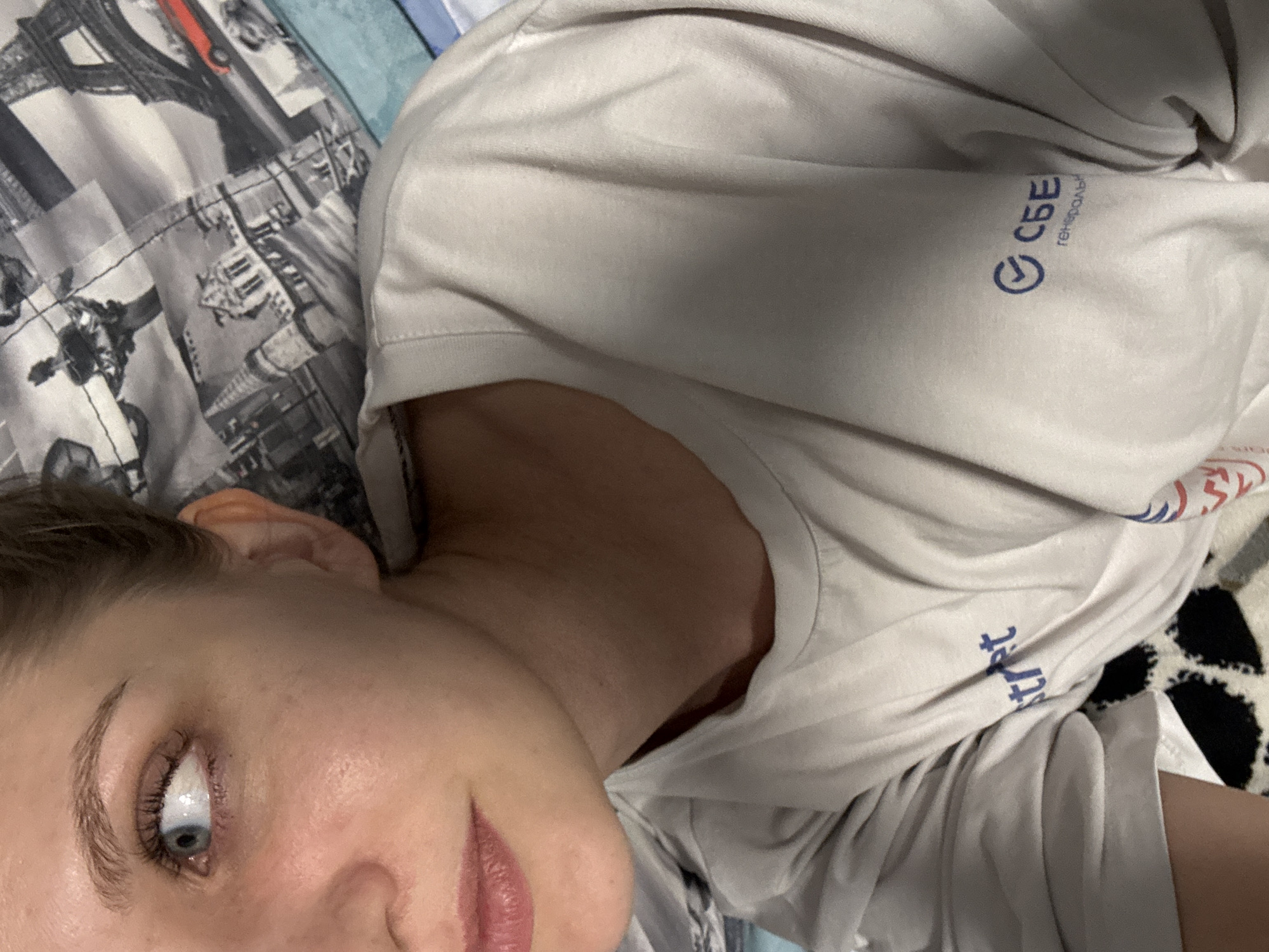 The image shows a close-up of a young woman's face and upper torso. She has fair skin, blue eyes, and light brown hair tied back. She is wearing a light-colored t-shirt with a logo that appears to be "СБЕ" in blue. The background shows a patterned fabric with what looks like a Paris theme, featuring the Eiffel Tower and other architectural elements. The lighting suggests an indoor setting, possibly a bedroom. There are no discernible elements in the image that would allow for the determination of her location within the city of Izhevsk, Russia.