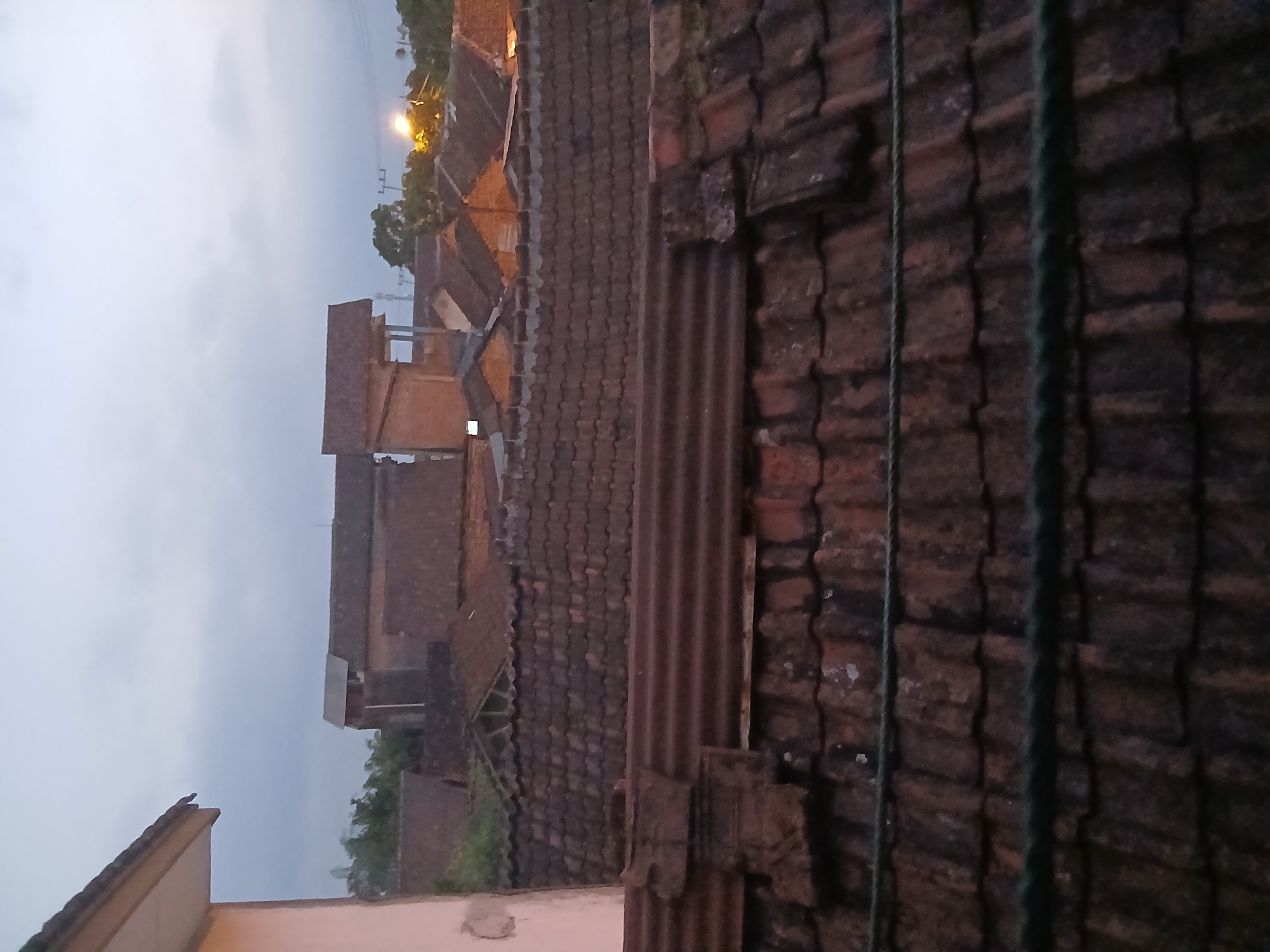 The image captures a view of rooftops in Kademangan Kulon, Indonesia, under a cloudy, twilight sky. The foreground is dominated by a series of weathered, reddish-brown tiled roofs, some showing signs of age and moss growth. A corrugated metal sheet is visible on one of the lower roofs, and a dark green rope runs horizontally across the bottom portion of the image. In the middle ground, more tiled roofs of various heights and designs are apparent, interspersed with glimpses of building walls constructed from brick or plaster. Some of these structures appear to be residential houses. In the background, further buildings with tiled roofs are visible, along with scattered trees. A utility pole with visible electrical wires can be seen on the right side of the image, and a bright, warm light, possibly a streetlamp or a light from a building, illuminates the distant treeline. The overall scene suggests a residential area at dusk or dawn.