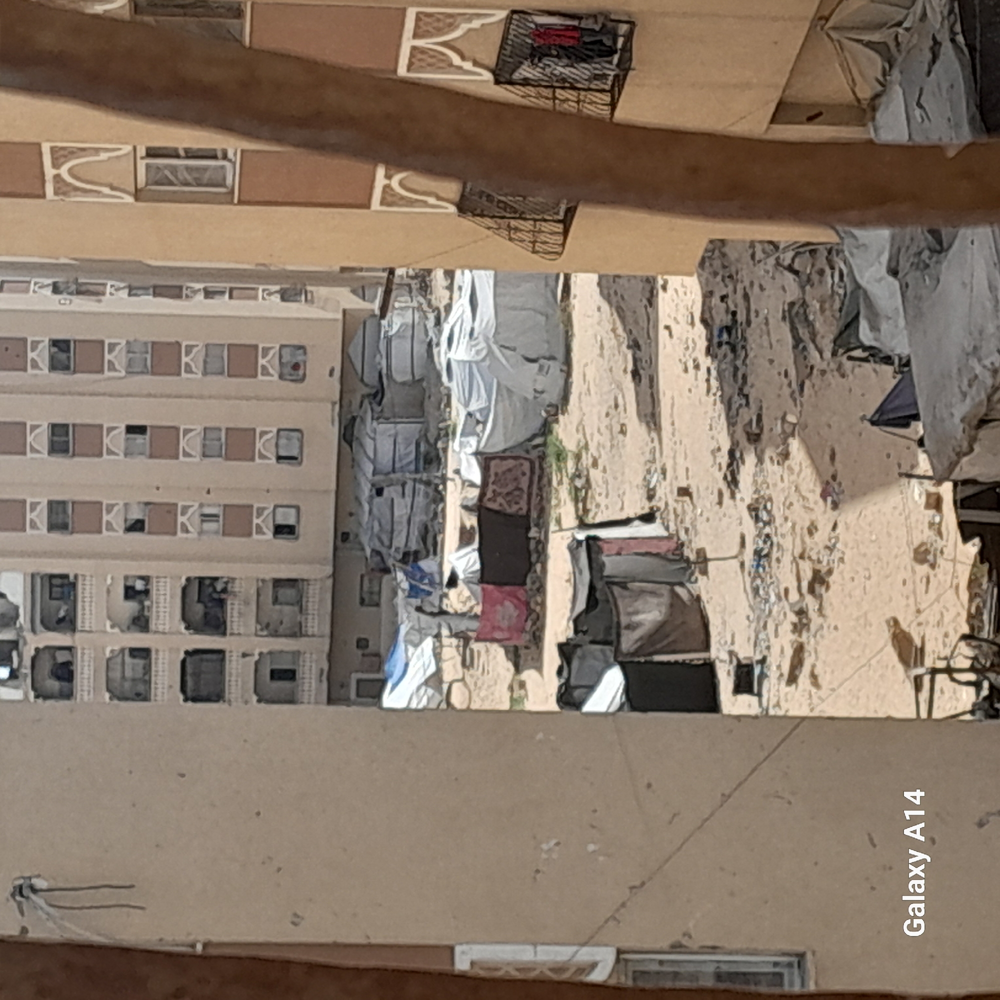 This photograph, taken in an unknown city, shows a view from above, looking down into a courtyard area. A tall apartment building is visible to the left, with numerous windows arranged in a grid-like pattern. The courtyard below is filled with what appear to be makeshift shelters or tents, constructed from white materials and other assorted fabrics. Rugs and various objects are scattered on the ground. The background is a sandy, barren landscape, possibly indicating a desert environment. A camera watermark "Galaxy A14" is visible in the bottom right corner of the image.