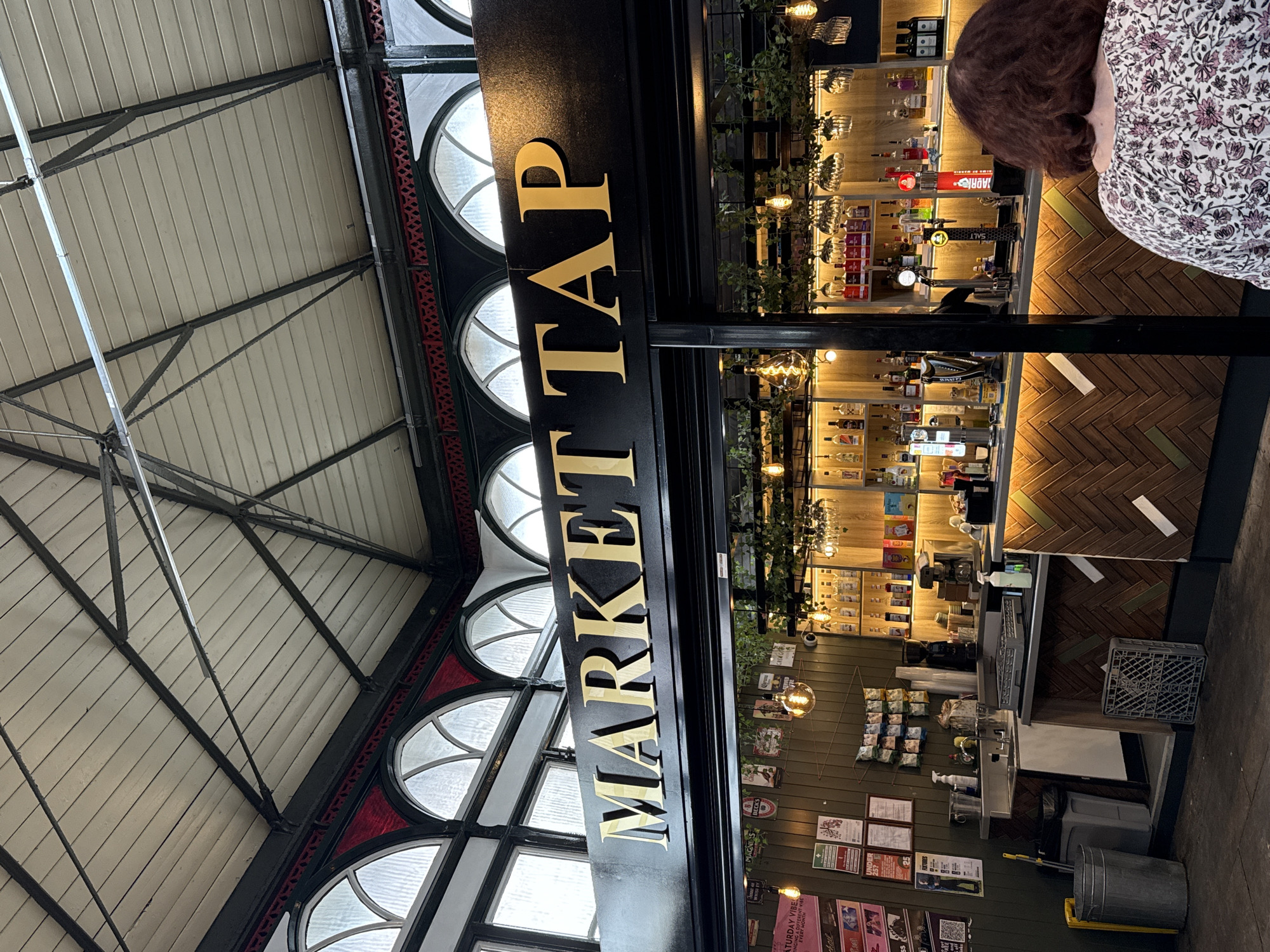 The image depicts the interior of a market taproom, likely located in Darlington, United Kingdom, given the context of the original image. The sign above the bar reads "MARKET TAP" in large, gold, capital letters on a black background. The taproom features a wooden counter with a chevron pattern and shelves stocked with various bottles of alcohol. Behind the bar, there is a coffee machine, sinks, and cleaning supplies. Pendants with Edison bulbs hang from the ceiling, casting a warm glow. In the foreground, a woman with curly brown hair is seen from behind, wearing a floral-patterned blouse. The ceiling is a vaulted structure with visible wooden beams and arched windows. The overall atmosphere suggests a lively market setting with a bar area.