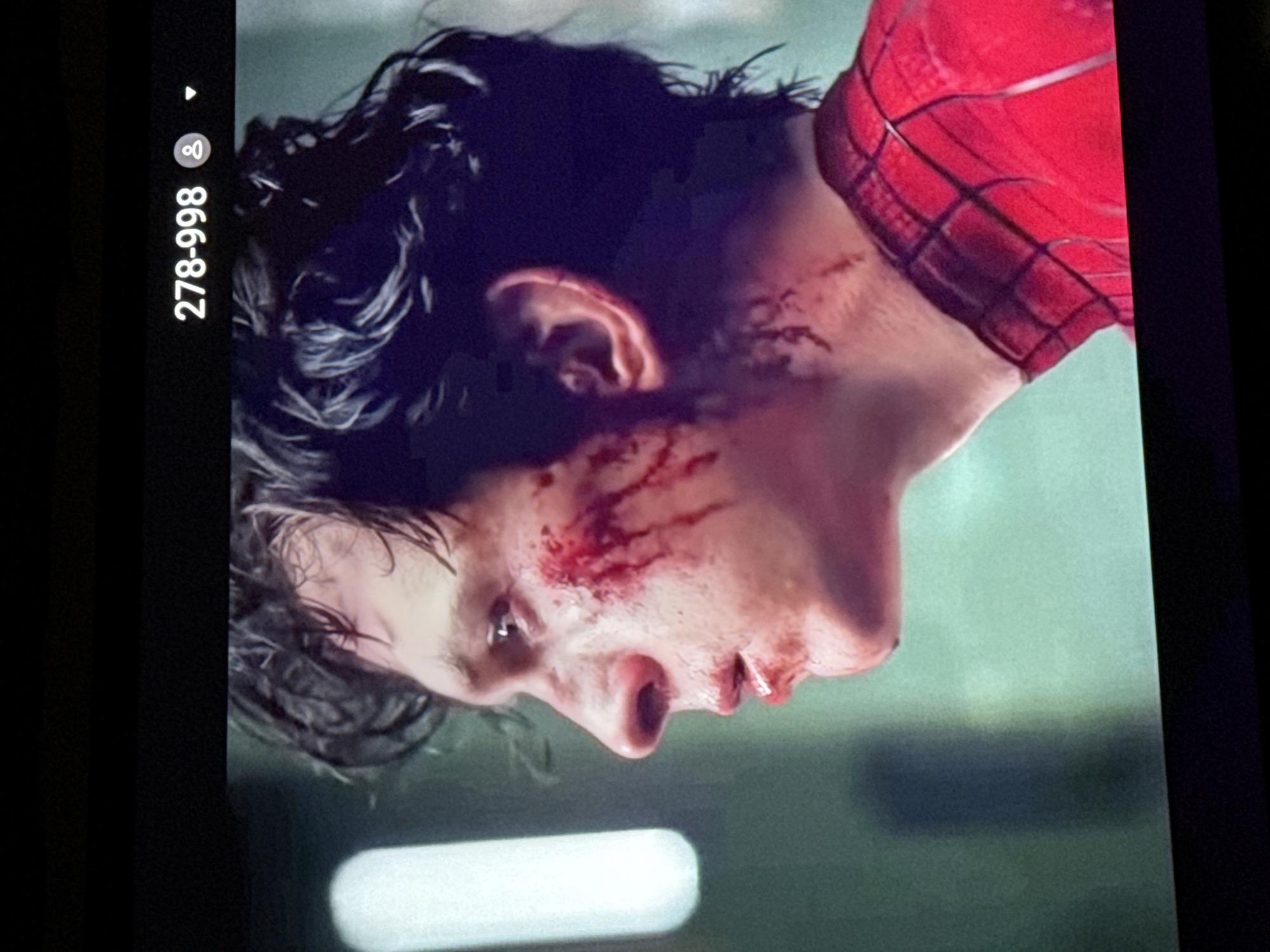 The image shows a close-up, side profile of a person wearing a red and black Spider-Man costume. The person's face is injured, with cuts and blood on their cheek and around their mouth. Their hair is dark and appears wet. The background is blurred, with a vertical white object on the left and a greenish-blue wall behind. There is also a digital overlay at the top of the image displaying "278-998" and a user icon. There is no indication of the city of Nyurba, Russia in the image.