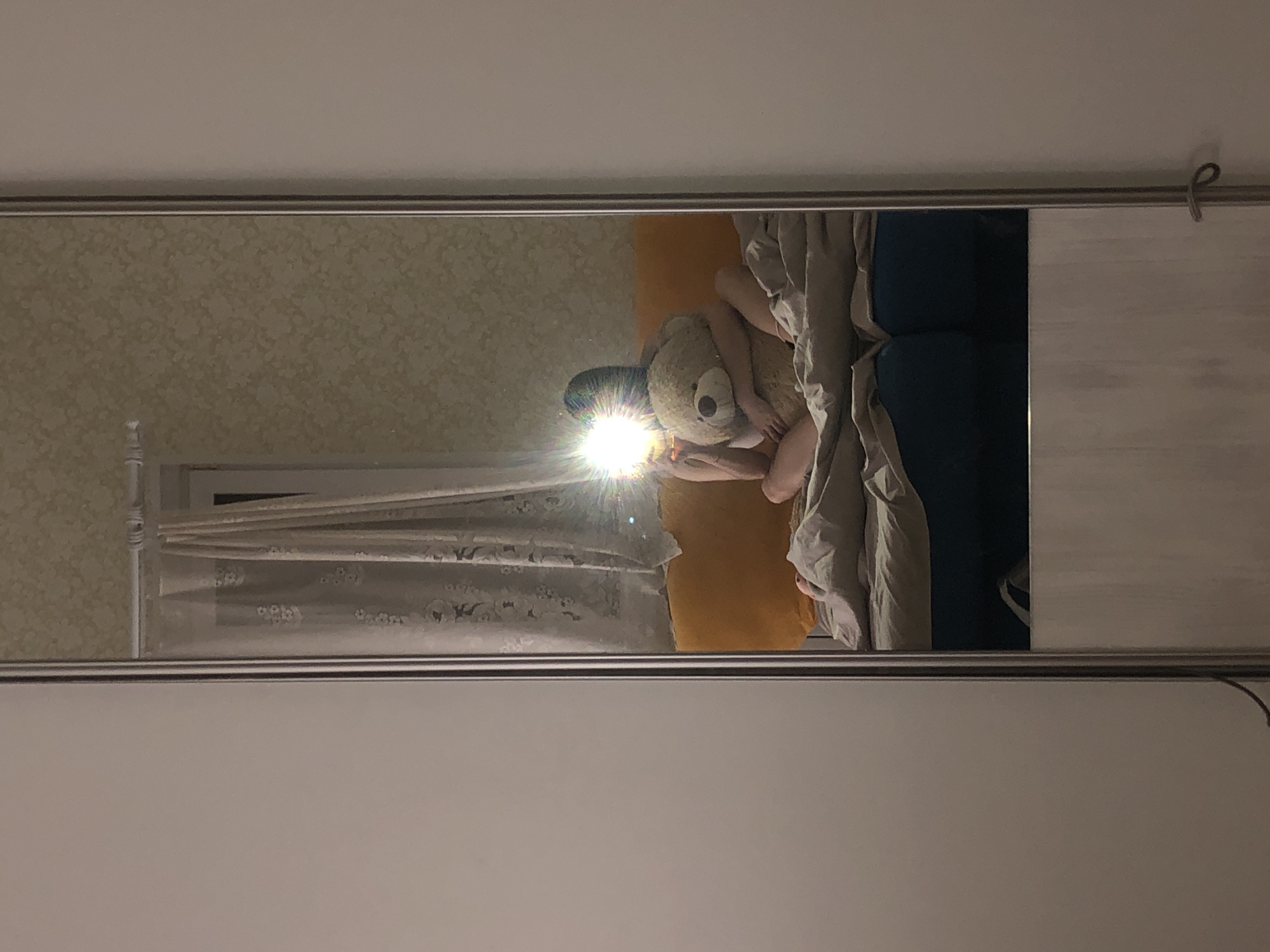 A reflection in a silver-framed mirror shows a person lying on a bed, cuddling a large teddy bear. The bed is made with blue and orange bedding, and a grey comforter is partially pulled over the person. A window with sheer, patterned curtains is visible in the reflection. The wall behind the bed has a textured, mottled wallpaper. The room itself, where the mirror is located, has a plain white wall. The city location of Noviy, Russia is not discernible.