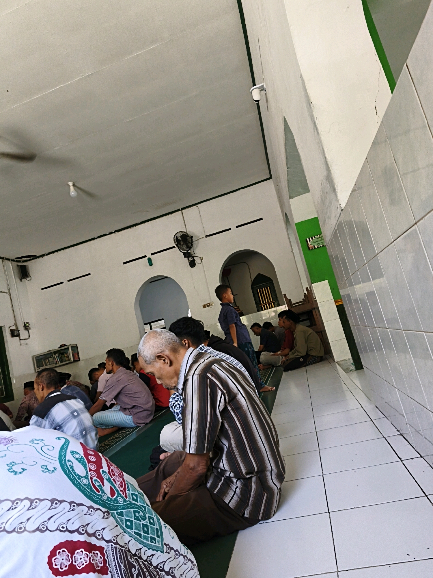 The image depicts the interior of a mosque in Desa Dibal, Indonesia, where several men are gathered, likely for prayer. The men are seated on a green carpet on the floor, facing forward. The architecture features white walls, a white tiled floor, and arched doorways. A ceiling fan is visible on the wall, along with a light fixture and some electrical wiring. There is a shelf on the wall containing books. The overall impression is one of a simple, functional religious space. The men are dressed in traditional clothing, and their postures suggest a state of reverence or contemplation.