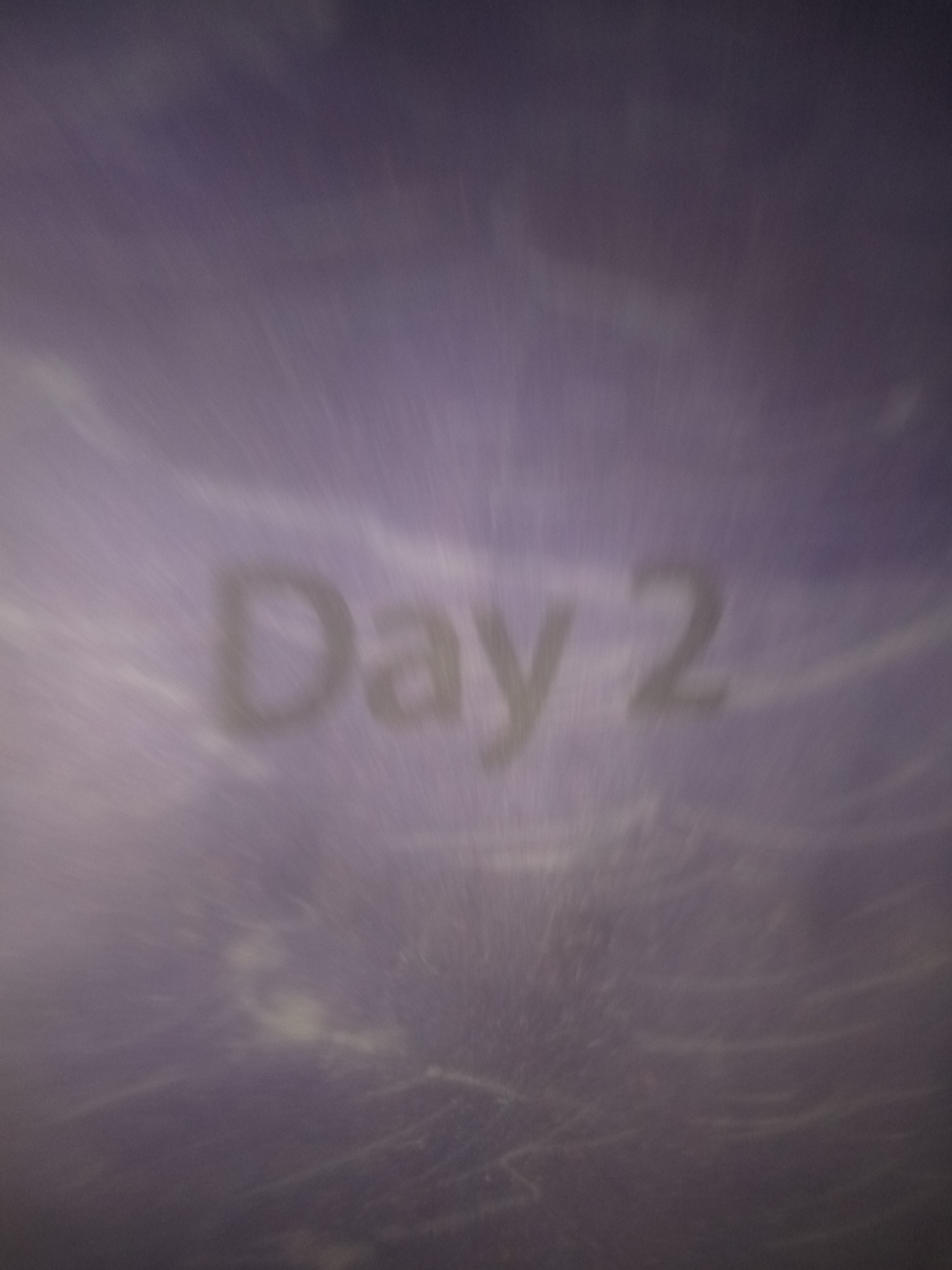 The image shows the text "Day 2" in blurry, light-colored letters against a dark purple, textured background. There are streaks and smudges on the surface, giving it a worn or aged appearance. The background appears to be a fabric or painted surface with some subtle lighter patches and streaks. There are no people or discernible objects within the scene. The image is too abstract and lacks specific details to determine a location within Jauro Yinu, Nigeria.