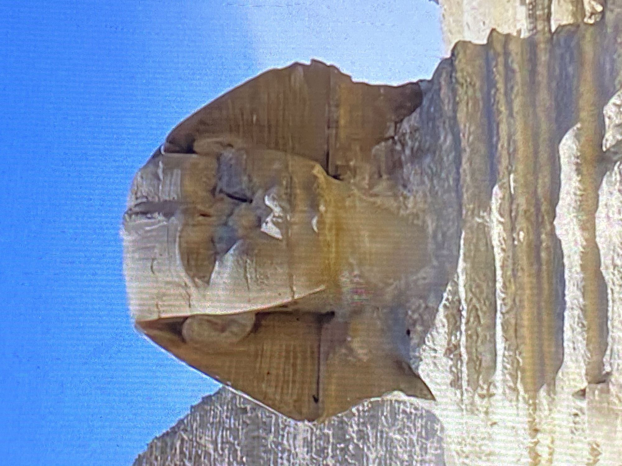 The image shows a close-up of a partially deteriorated stone structure against a clear blue sky. The structure appears to be an architectural fragment with weathered surfaces and eroded details. Based on the location, it is likely a section of the Bolívar Bridge in Tinaquillo, Venezuela. The stone has a textured surface with vertical striations and is colored in shades of brown and beige. Parts of the structure are missing or broken.