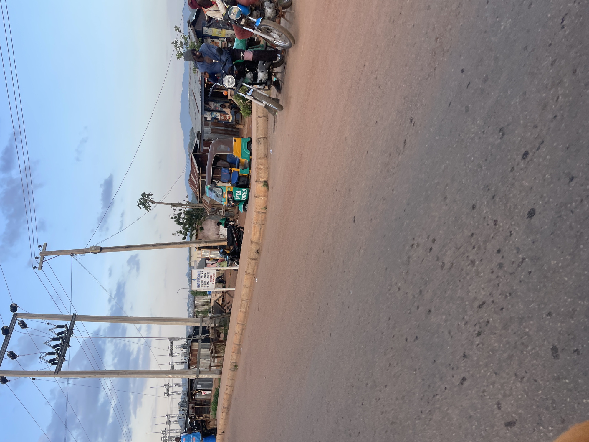 The image captures a vibrant street scene in Nabatum, Nigeria, likely during the late afternoon or early evening under a light blue sky with scattered clouds. The primary focus is a paved road that curves slightly to the left, flanked by an unpaved shoulder and various structures.
On the right side of the road, several individuals are gathered near and on motorcycles. One man, wearing a blue shirt and a dark cap, is seated on a motorcycle parked on the shoulder. Another motorcycle is visible directly behind him. Further along, numerous informal shops or stalls with corrugated metal roofs line the roadside, indicating a commercial area.
On the left side of the road, a yellow and green trishaw (motorized tricycle taxi) with the license plate "PTA-0285" is prominently parked. Overhead, multiple utility poles with complex wiring stretch across the frame, carrying power lines. In the background, beyond the immediate structures, distant mountains or hills are visible under the soft light.
A white sign, partially obscured and rotated, is visible on the right side of the road near the utility poles. Text on the sign includes "SOLANTO GROUP OF COMPANIES CORPORATION" and further details such as "P.O BOX", and potentially locations like "KADUNA" and "ABUJA", suggesting business operations. The asphalt road surface shows visible texture and speckles. The scene depicts typical daily life and commerce in a Nigerian urban or semi-urban setting, characterized by local transportation, informal businesses, and utility infrastructure.