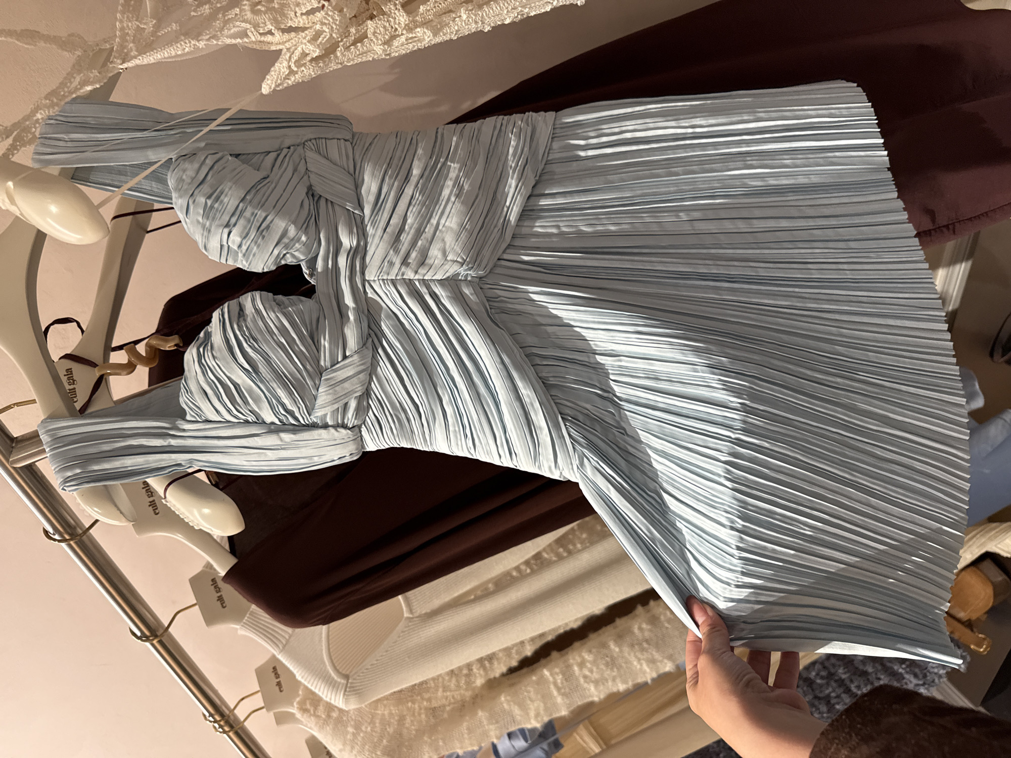 A blue pleated dress hangs on a clothing rack among other garments. The rack is located in an unidentified retail environment in the city of Sunrise, United States of America. The garments on the rack include a brown top, a white sweater, and a lacy white garment. A hand is seen holding up the edge of the blue dress. The hangers are marked with "cult gaia".