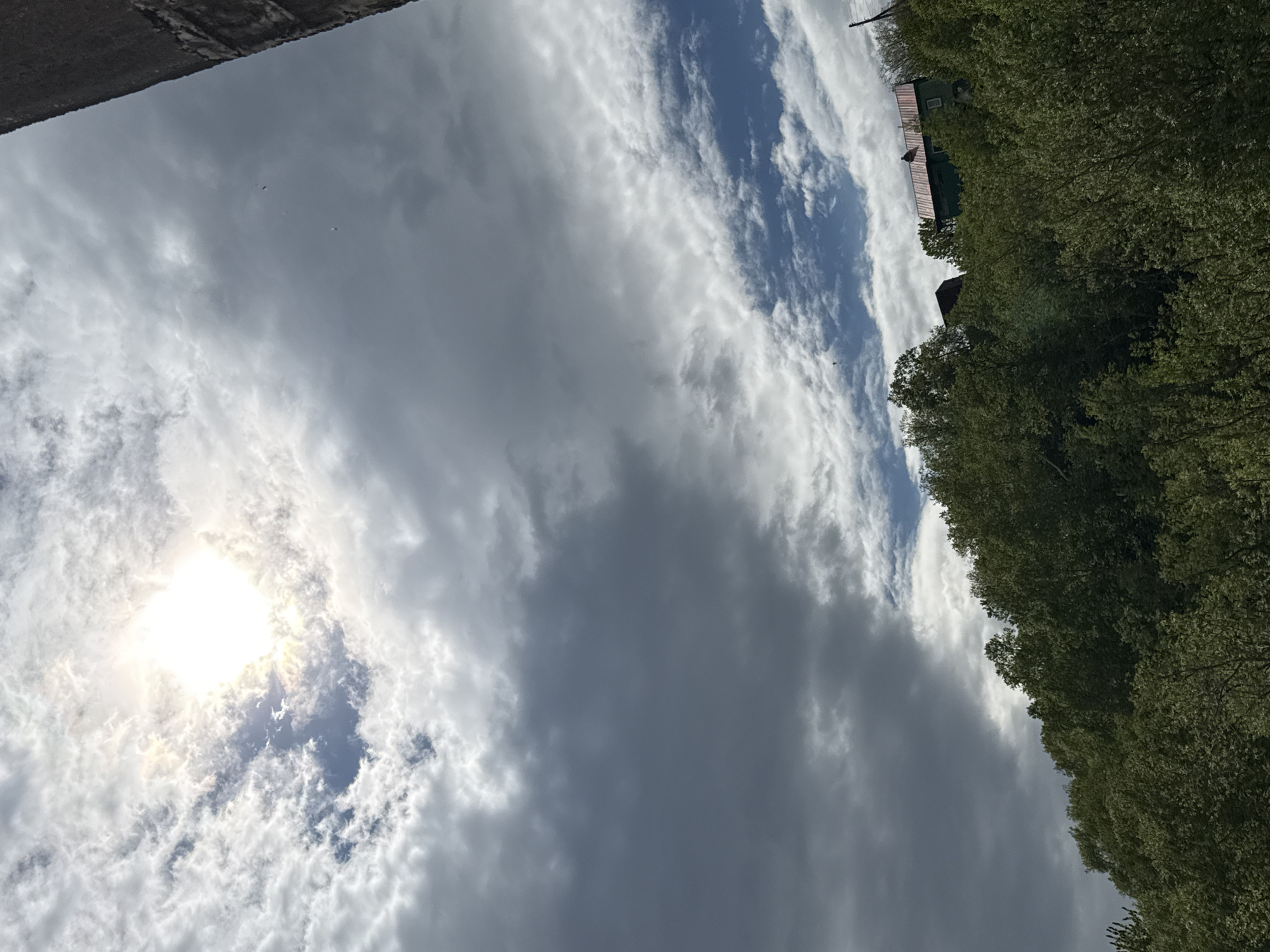 The image shows a view of a green hillside covered in lush trees. On top of the hill, two buildings are visible. The larger building has a red metal roof and green walls, with some visible windows. To its left, a smaller, darker building with a visible roofline is partially obscured by trees. The sky above is partly cloudy with the sun shining through a break in the clouds, creating a bright, ethereal glow. A dark, vertical structure, possibly a concrete pillar or wall, is visible on the far right side of the image, extending downwards from the top. The scene appears to be taken from a low vantage point, looking upwards towards the hill and sky. The environment suggests a natural, possibly rural or suburban setting on the outskirts of a city.