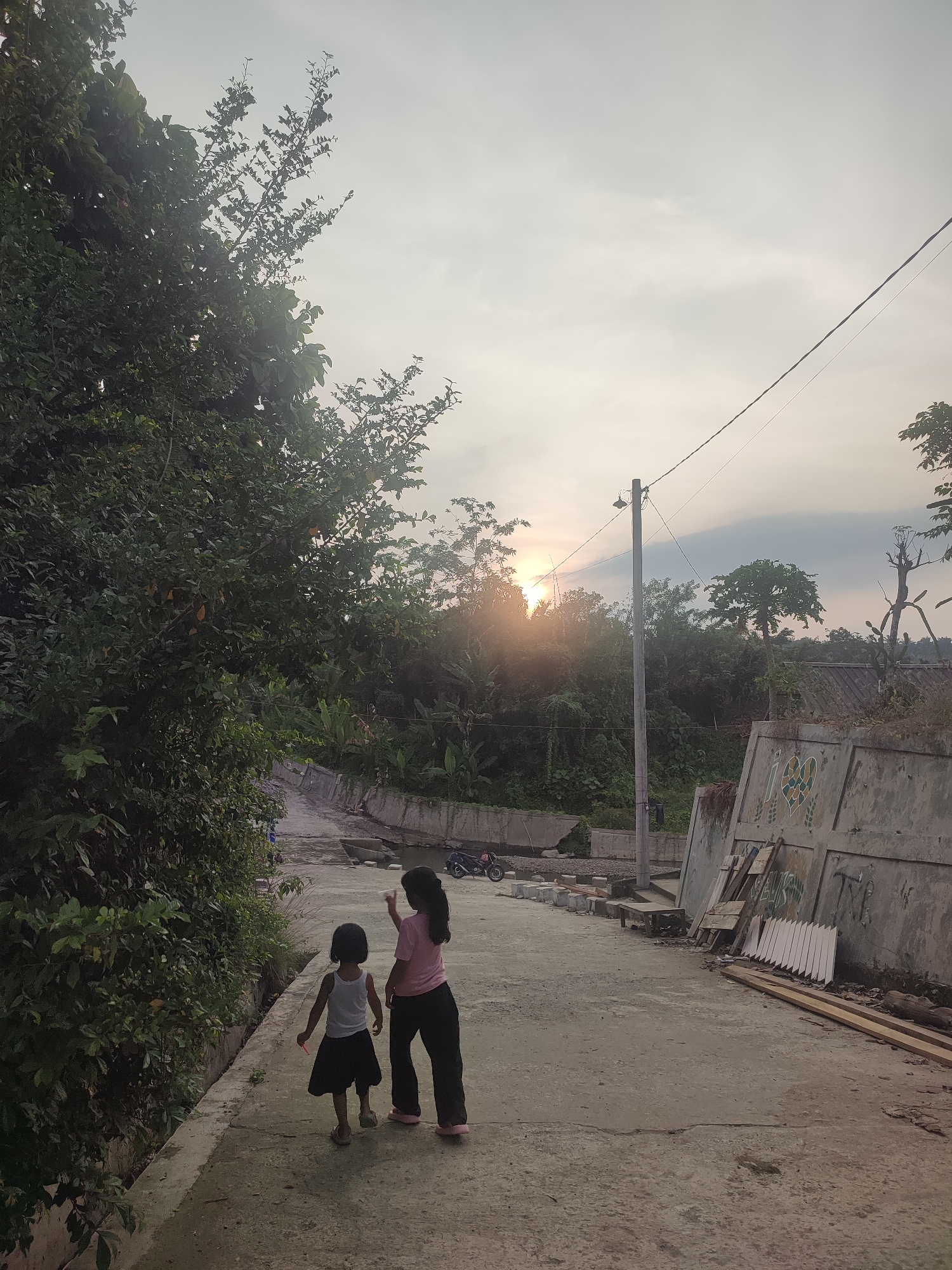 Two children, a younger girl and an older girl, are walking on a paved road in what appears to be a residential area in Saweuk, Indonesia. The younger girl is wearing a white tank top and a dark skirt, while the older girl is wearing a pink shirt and dark pants. The older girl is pointing towards the horizon, where the sun is setting behind trees. Lush greenery flanks the left side of the road, and a concrete wall with graffiti and stacked building materials lines the right side. A utility pole stands upright along the road, and a motorcycle is parked in the distance. The sky is partly cloudy with the sun creating a bright glow. A stream of water runs along the side of the road.