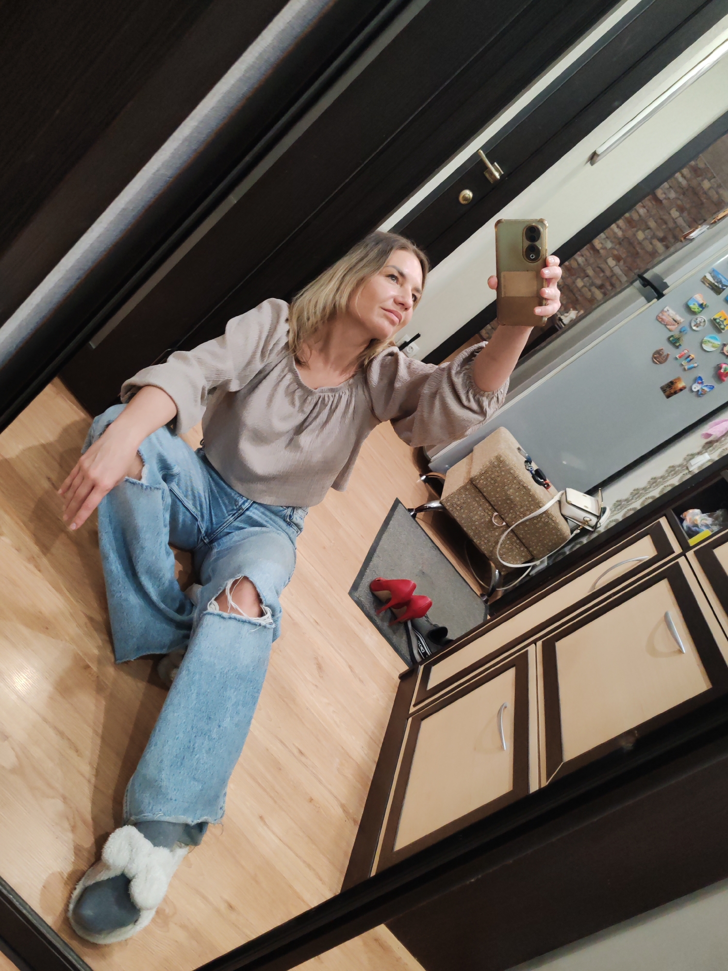 A woman is taking a selfie while sitting on a wooden floor indoors. She is wearing a light brown blouse, ripped blue jeans, and grey slippers with white fur trim. She is holding a cell phone with a green case in her right hand. A doorway with dark brown trim is in the upper left corner of the image. Behind the woman is a kitchen area with a light grey refrigerator covered with magnets, and light tan cabinets with dark brown trim. A beige patterned bag sits on a chair behind her. Red high-heeled shoes are visible on a grey doormat. It is impossible to verify the city or location with the provided information.