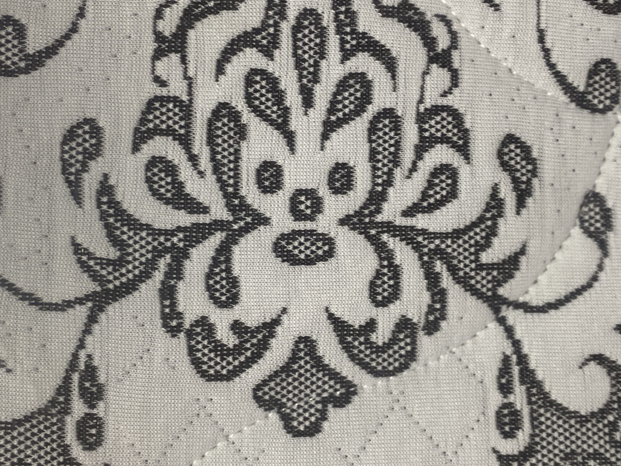 The image is a close-up, overhead view of a textured fabric with a damask-style pattern in black and off-white. The fabric appears to be a quilted material, with a subtle diamond-shaped stitching pattern visible beneath the printed design. The black pattern consists of intricate floral motifs, including swirling leaves and petal-like shapes, some filled with a dense, cross-hatched texture and others with solid black areas. The off-white background fabric has a visible weave and numerous tiny perforations, suggesting it might be a breathable material. The lighting is even, with no harsh shadows, indicating diffused light. There are no people, animals, or specific landmarks visible in this tightly cropped image. Without further information or context, it is impossible to determine the location in Novokuznetsk, Russia.