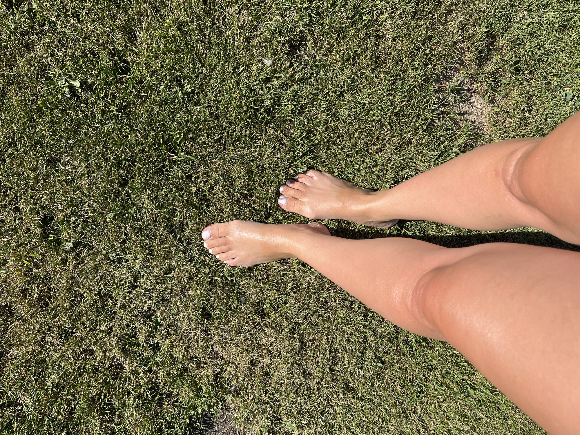 The image shows a pair of tanned, bare legs standing on a patchy, dry-looking green lawn. The left leg is closer to the camera and its knee is bent. The toes of both feet are visible, with the toenails of the left foot painted white and the right foot having some white nail polish on the big toe. The lighting suggests it is daytime, with a clear sky casting shadows. The environment appears to be an outdoor grassy area, possibly a park or backyard. There is no indication of the specific location in Novosibirsk, Russia.