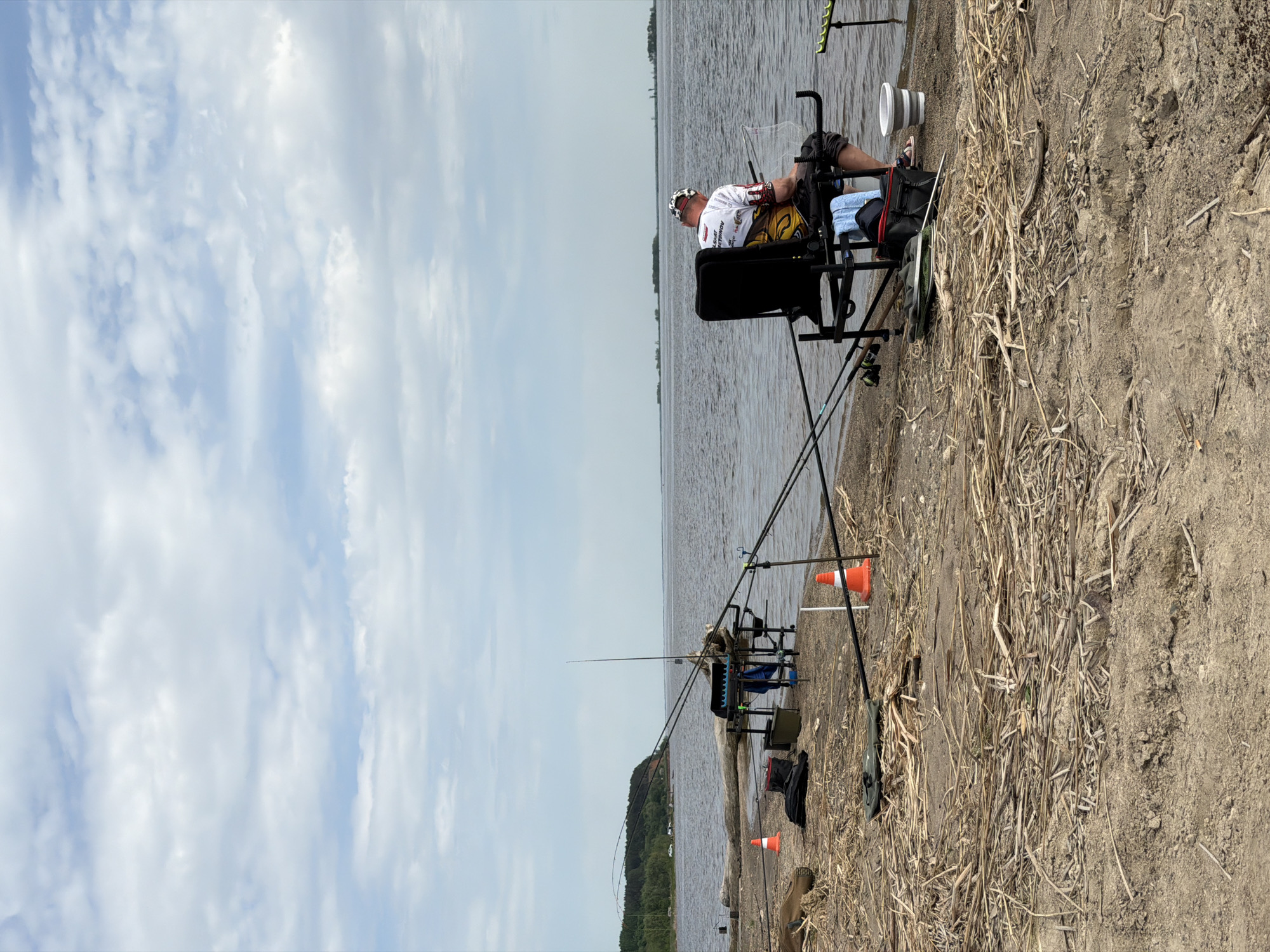 The image shows a man fishing on the edge of a body of water. He is seated in a specialized fishing chair with equipment, including multiple fishing rods set up along the bank. The ground consists of dirt and dry reeds or grass. The water is calm, and the sky is partly cloudy. The scene appears to be in Krasnyi Bor, Russia, based on the information given.