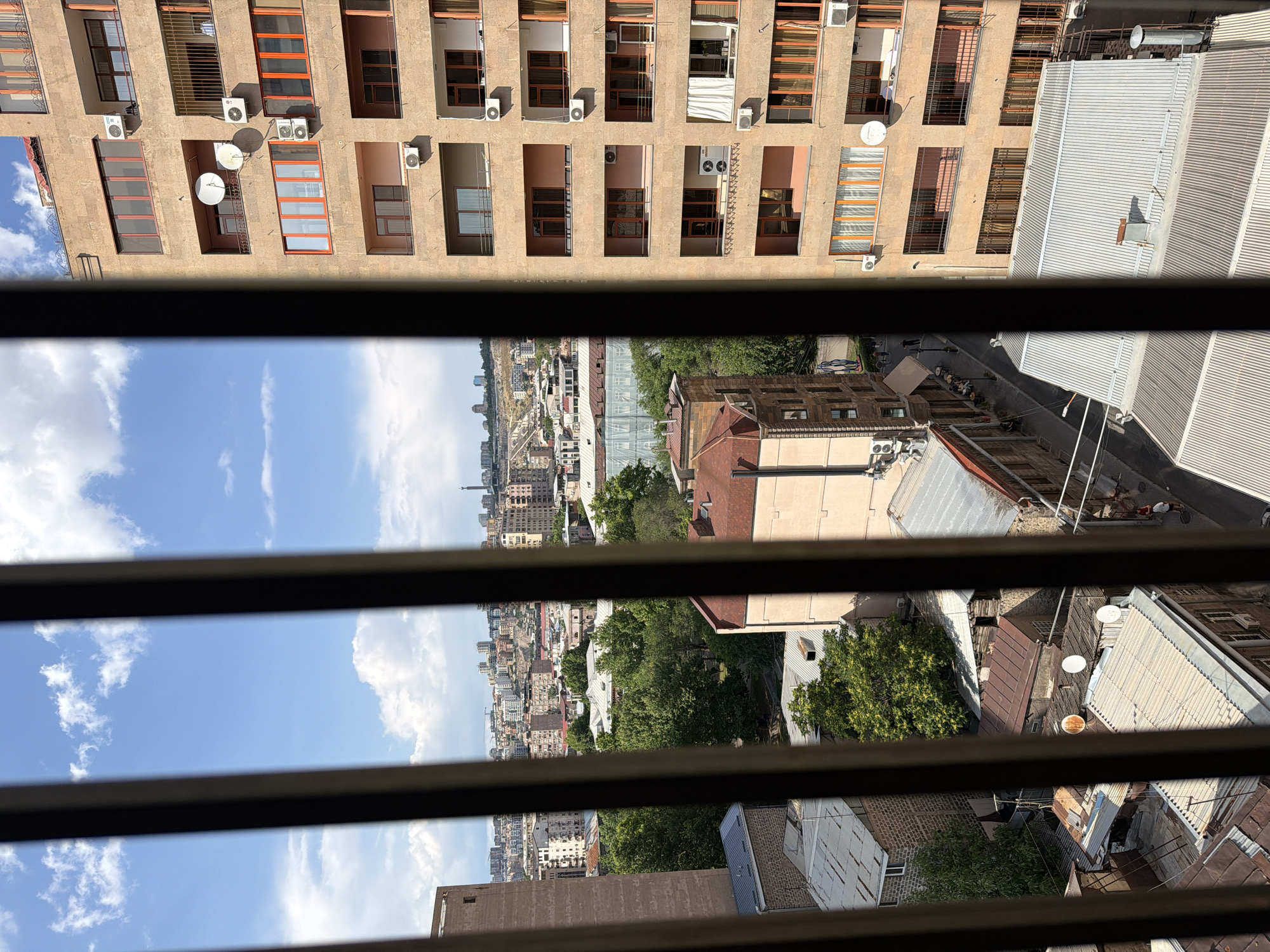 The image captures a view of a cityscape through vertical bars, likely from a window or balcony. The scene is set in Yerevan, Armenia, as evidenced by the distinctive architecture. In the foreground, to the right, is a tall, beige apartment building with multiple floors. Each floor features several windows with orange frames, and air conditioning units are visible on some of the exterior walls. Several satellite dishes are mounted on the building's facade.
To the left of this building, and further back, are smaller, older buildings with rust-colored, corrugated metal roofs. These structures appear to be residential or commercial spaces. Beyond these, a dense urban landscape unfolds, with numerous buildings of varying heights and styles stretching towards the horizon. The city is built on a hilly terrain, with some structures perched on inclines. Lush green trees are interspersed throughout the urban environment, particularly in the mid-ground.
In the distance, the sky is a bright blue, dotted with fluffy white clouds. The overall impression is of a sunny day in a moderately dense urban area. A narrow street is visible between the apartment building and the smaller structures, with a few people walking or seated at tables outside a cafe. The perspective is from an elevated position, looking down and across the city.
