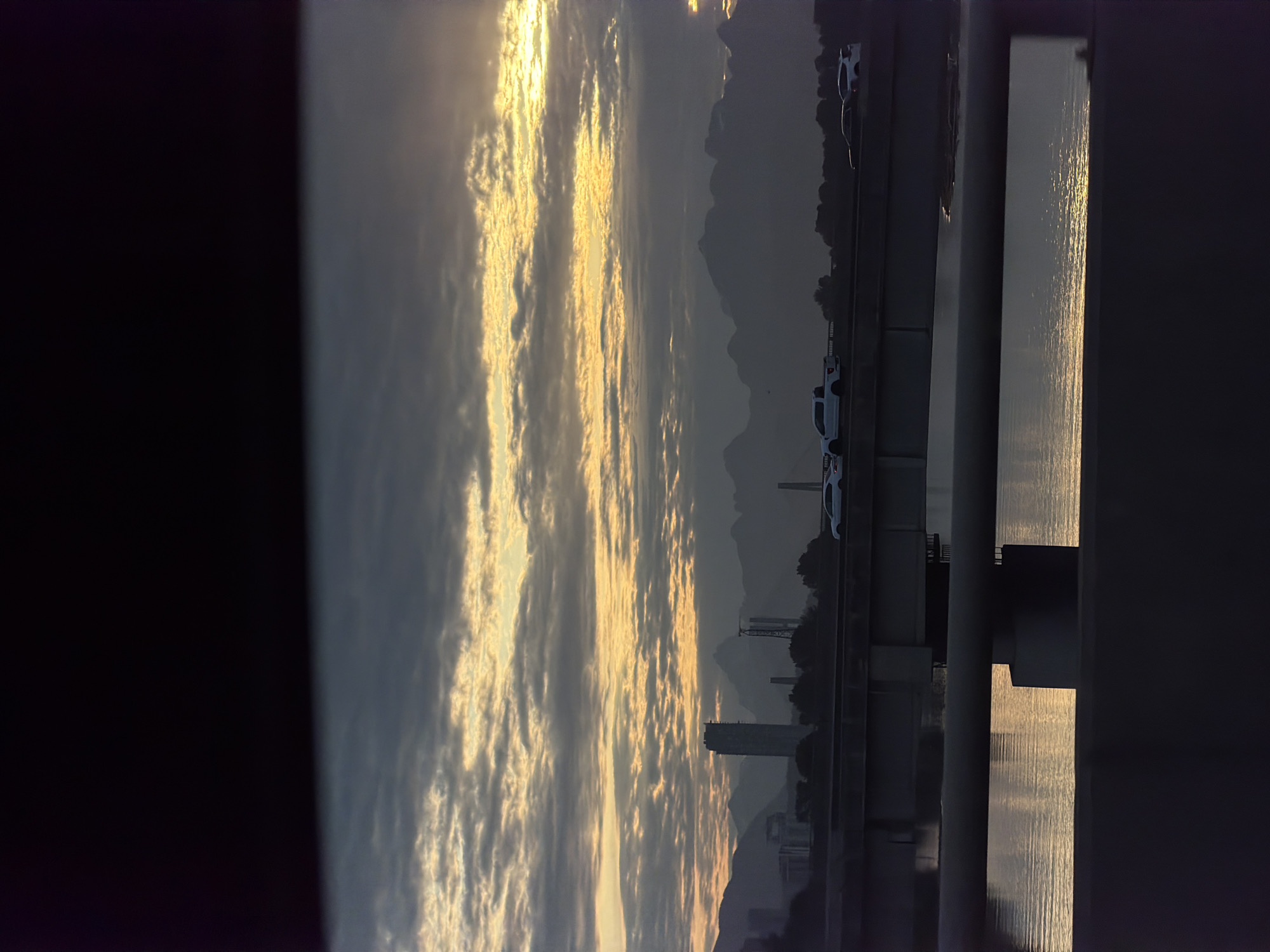 The image shows a dusky scene in Delta, Canada, likely at sunset. The view is framed by dark structures, possibly a bridge or building, on the left and right sides. In the center, a body of water reflects the golden light of the setting sun, creating shimmering patterns on the surface. Beyond the water, silhouetted mountains rise in the distance, adding depth to the landscape. A line of cars can be seen, indicating a road or bridge. In the middle distance, building-like structures stand out in the cityscape. Overall, the scene conveys a sense of tranquility and beauty in the Canadian landscape.