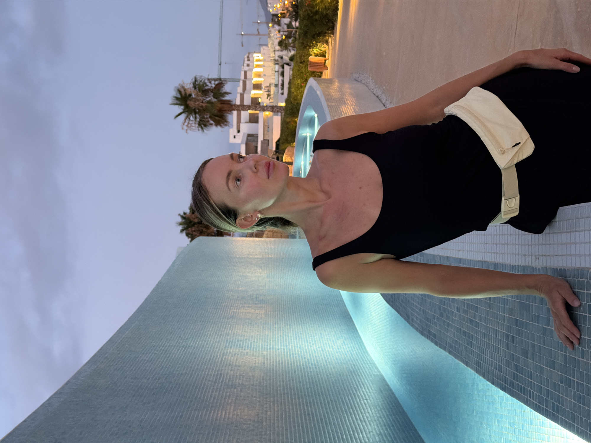 A woman is posing for a photo by a curved, illuminated pool at dusk in Finestrat, Spain. She is wearing a black dress with a cream fanny pack around her waist. The background features modern white buildings, palm trees, and construction cranes, suggesting a developing resort area. The sky is a dusky blue, indicating twilight. The woman is looking to her left, with her left hand resting on the tiled edge of the pool. The overall scene is serene and hints at a vacation setting.