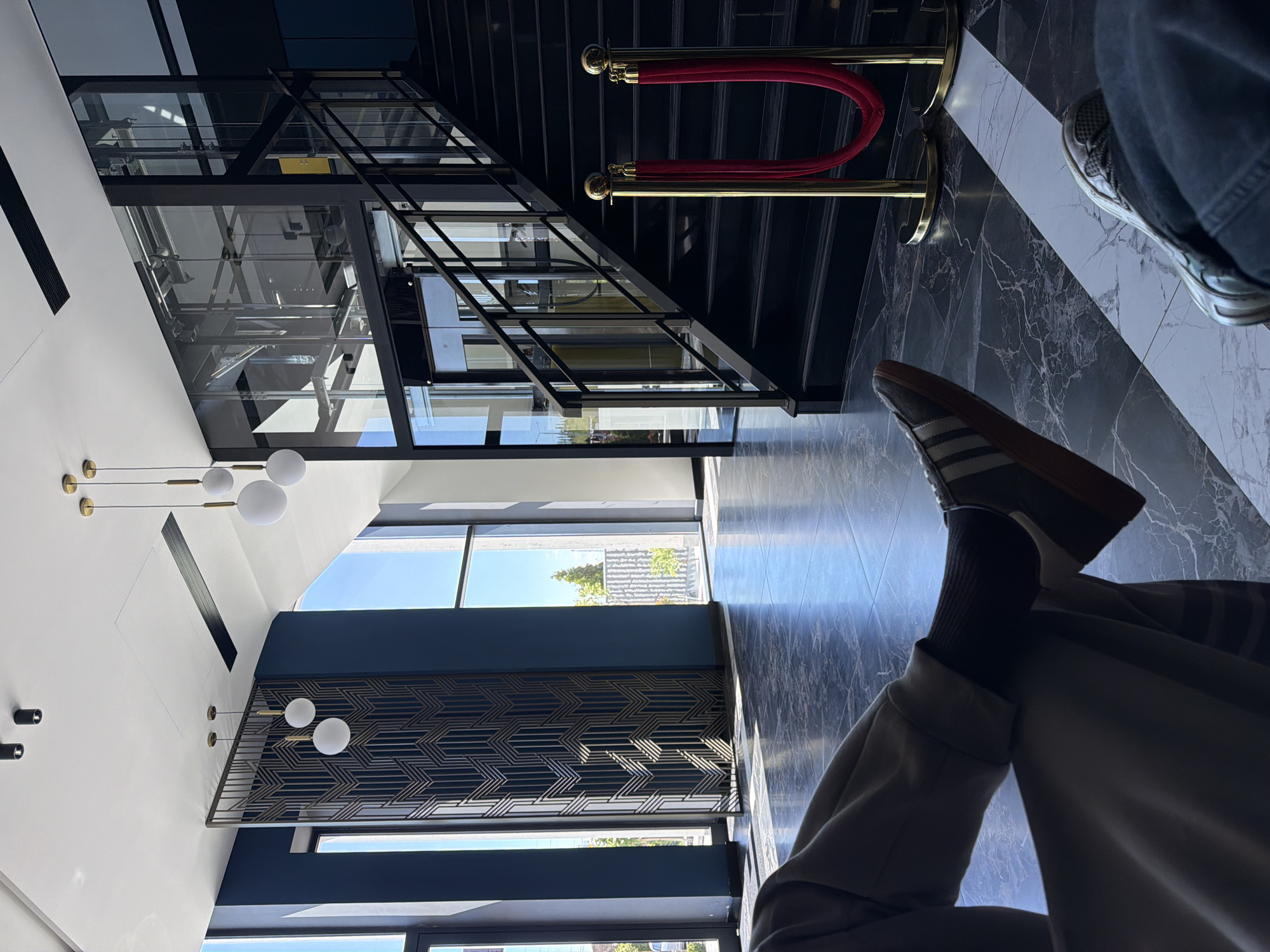 The image shows an interior view, apparently within a modern building located in Verin Ptghni, Armenia. The space features a contemporary design with a high ceiling, accented by modern globe lighting fixtures. A decorative geometric panel is on the wall. There is a glass-enclosed elevator shaft. A staircase is present, demarcated by gold stanchions and red velvet ropes. The flooring has a marble-like pattern in dark and light tones. A pair of legs, one crossed over the other, is visible, with a sneaker and a brown-soled shoe on the feet. A person is sitting in the foreground. A partial view through windows reveals a glimpse of greenery.