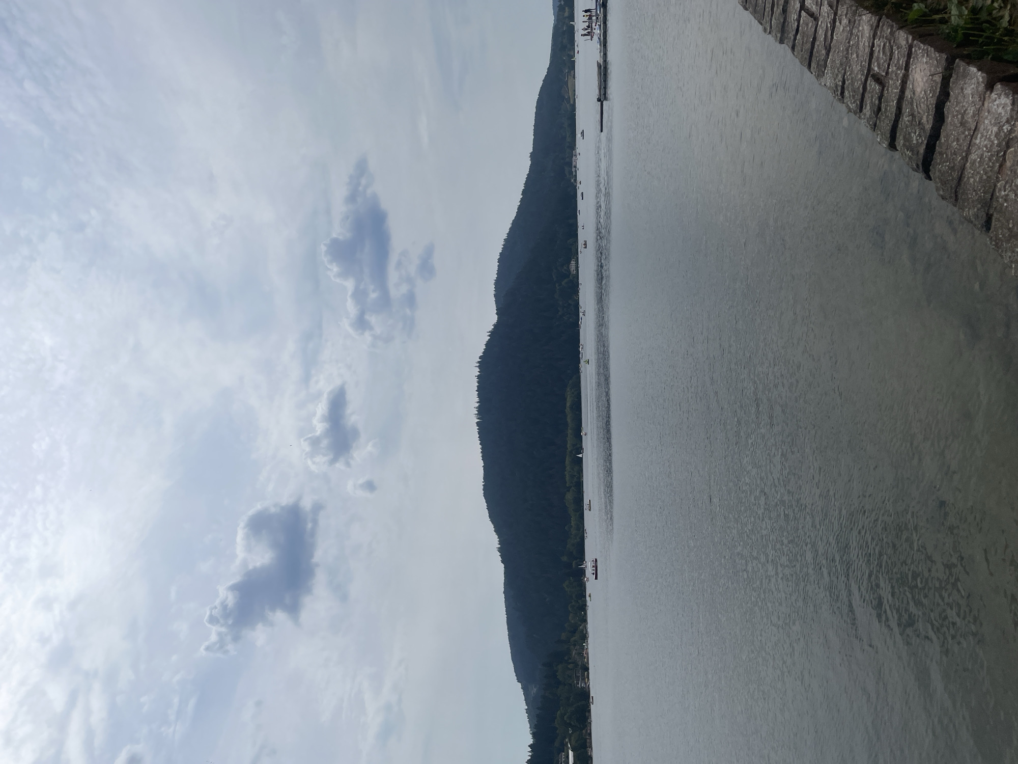 The image depicts a wide, calm lake with a dark, forested hill in the background. Several sailboats are scattered across the water, with some appearing to be docked or anchored near a pier on the right side of the frame. The sky is overcast with scattered clouds. A stone embankment lines the lower right corner of the image, with some green vegetation growing along its edge. The scene appears to be a tranquil, scenic view of a lake and its surrounding landscape in Gérardmer, France.