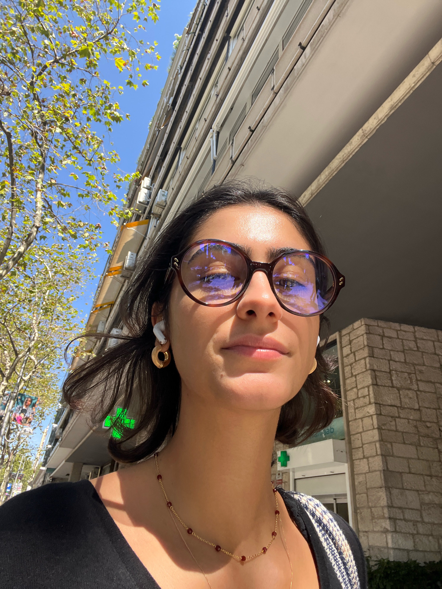 A close-up, low-angle, selfie-style shot captures a young woman with dark hair, wearing large, round, tortoiseshell-patterned glasses and white wireless earbuds. She is adorned with a gold necklace featuring small red beads and a circular gold earring on her left ear. She is wearing a black shirt and a jacket with a striped pattern over her right shoulder. The background shows a modern, multi-story building with a beige facade, accented by stone-brickwork at the lower right. To the left of the building, a leafy green tree with a bright blue sky above provides a natural element. A pharmacy sign with a green cross is visible on the building's exterior. The overall scene suggests an urban setting, possibly during daylight hours, with a clear sky. The reflection in the woman's glasses indicates the presence of windows and possibly other buildings or a sky reflection. The image does not contain explicit information about the specific location in Madrid, Spain, beyond its urban characteristics.