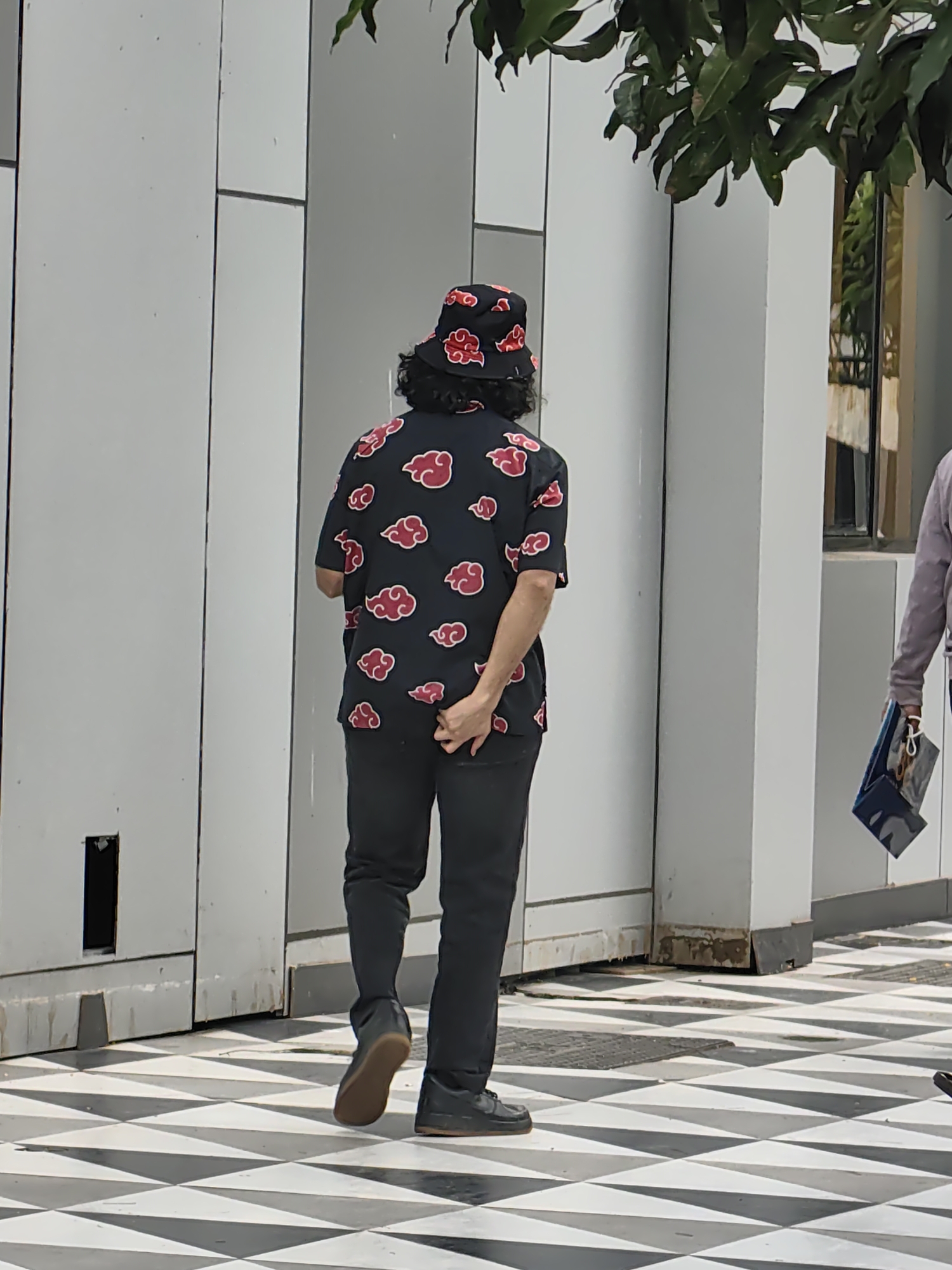 A person wearing a black bucket hat and a black short-sleeved shirt with a red cloud pattern is walking away from the camera on a checkered tile sidewalk. The person's right hand is behind their back, tucked into their pants. They are also wearing black pants and dark shoes. In the background, there is a white wall with vertical panels and some green foliage hanging from the top of the image. To the right of the person, another individual is walking away from the camera, carrying a blue bag. The scene appears to be in an urban environment, possibly a commercial area in Mumbai, India, given the architectural style and the checkered tiling which is common in certain parts of the city.