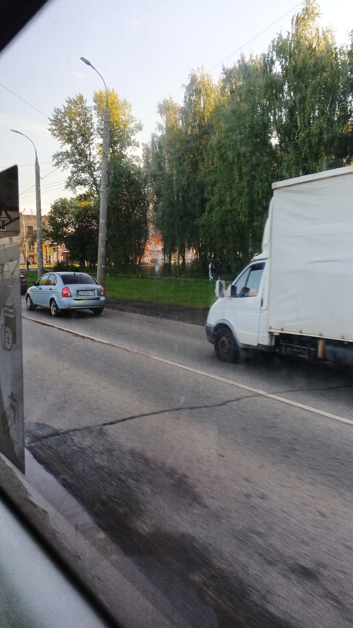 The image depicts a street scene in Izhevsk, Russia. A light blue sedan is parked on the side of the road, and a white truck is driving past. Trees line the street, and a building can be seen in the background. The sky is clear and the sun is shining.
