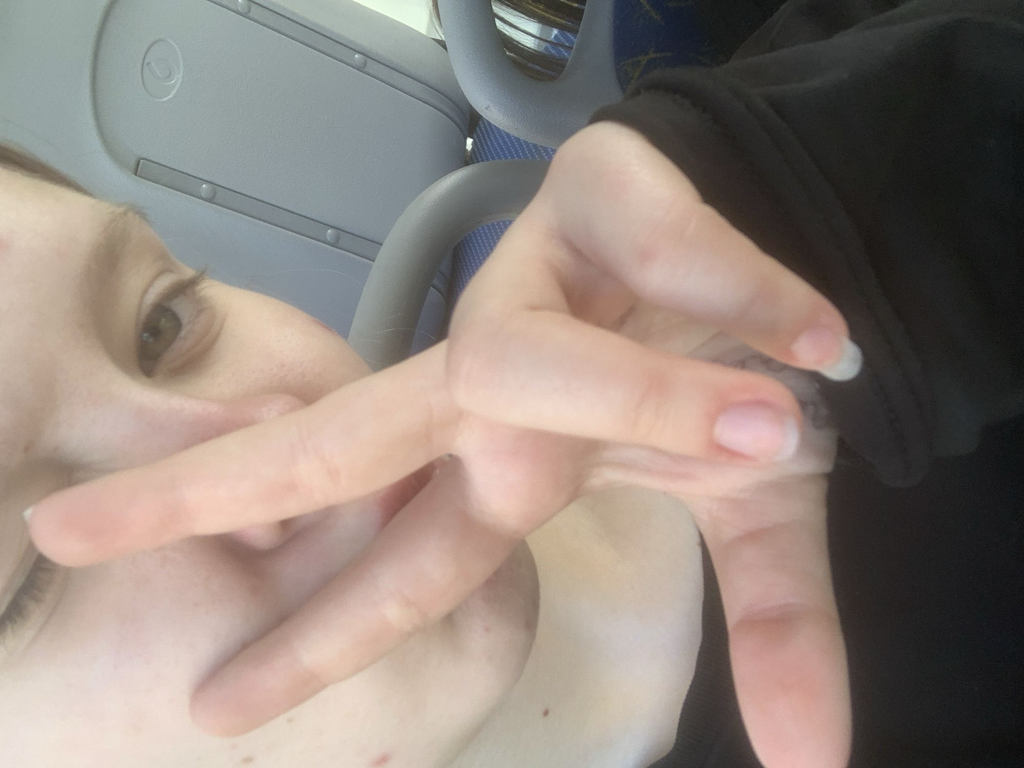 The media file consists of an original image and several cropped sections, depicting a person taking a close-up selfie inside a public transport vehicle.
**Subjects:** The primary subject is a person, whose face is partially visible and obscured by their right hand. The visible part of their face includes an eye (likely brown), part of the nose, and a cheek, suggesting light skin. The hand is prominent, making a "V" or peace sign with two fingers extended upwards. A black garment, possibly a sleeve or jacket, is visible on the person's left side.
**Objects:** The background elements clearly indicate the interior of a vehicle. These include light grey overhead panels featuring a small circular graphic symbol (resembling a stylized 'C' or hook within a circle) and two parallel grey strips with small, round rivets. Further back, two grey, curved handrails are visible, typical of bus or train interiors. Behind these, blue upholstery with a light geometric pattern can be seen. The person's fingernails are short and light-colored, with one appearing to have a white tip.
**Scene/Setting:** The image captures a moment inside a public transport vehicle, such as a bus or train, during what appears to be daytime. The person is holding the camera at an angle, resulting in a tilted perspective.
**Activity or event taking place:** The person is actively taking a selfie, using their hand to make a peace sign, partially covering their face in the process.
**Notable details:** The light conditions suggest it is daytime. The vehicle interior appears functional and somewhat utilitarian, typical of public transport. There are no specific weather indicators. No clear, legible text is visible within the image. The person's expression is not fully discernible due to the obscured face and selfie angle, but the hand gesture typically conveys a casual or friendly sentiment.