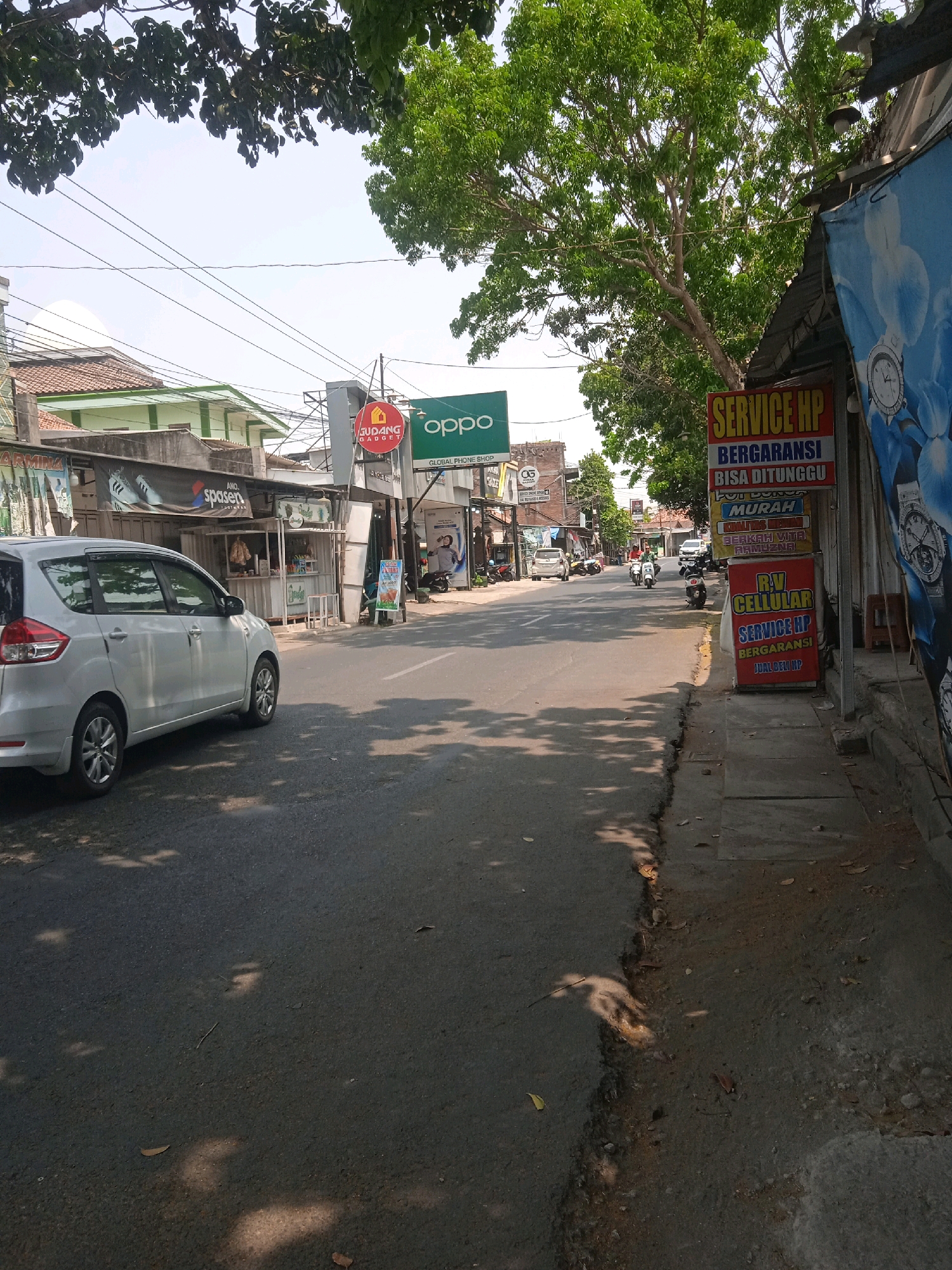 The image shows a street scene in Bintoro, Indonesia, on a sunny day. A silver minivan is parked on the left side of the road, with the left side of the vehicle visible. The road is paved and appears to be a two-lane street. On the right side of the street, there are several storefronts. One storefront has a large sign with blue and white text that reads "SERVICE HP BERGARANSI BISA DITUNGGU MURAH". Below this sign is another sign for "R.V CELLULAR SERVICE HP". Further down the street, on the left, is a small food stall with customers. Trees with green foliage hang over the street from the left and right sides. Several power lines are visible crisscrossing the sky. The overall impression is a typical street scene in a commercial area of an Indonesian town.