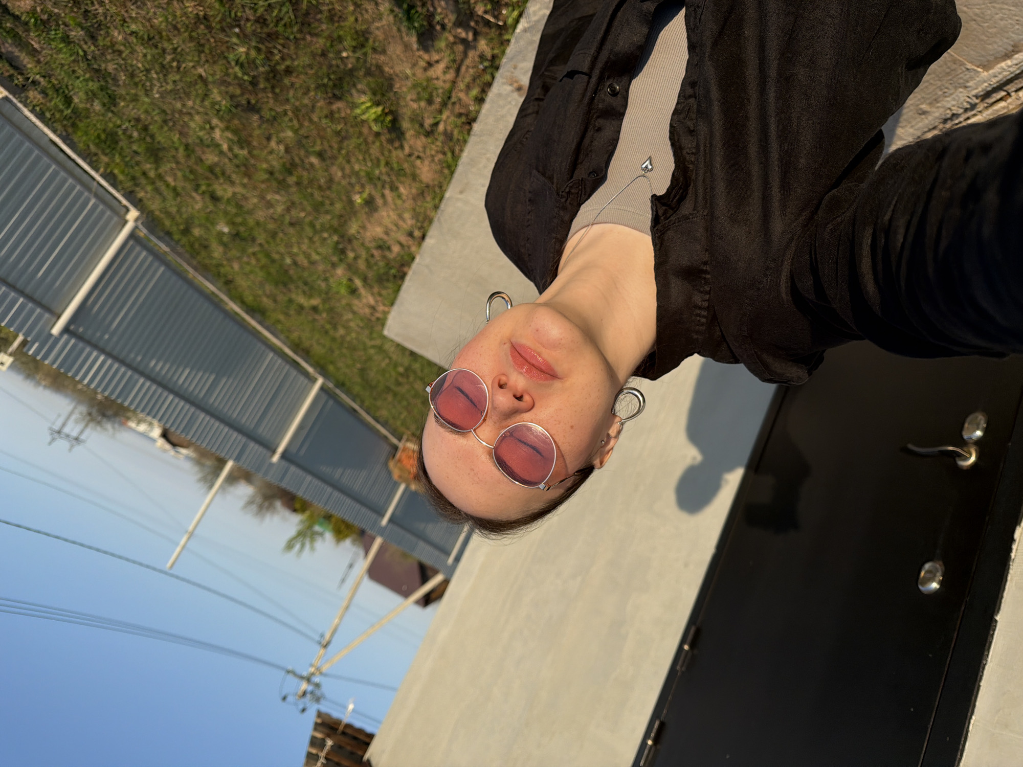 A person wearing pink-tinted round glasses, large hoop earrings, and a dark button-down shirt over a light-colored top is posing for a selfie. The person is standing in front of a light gray or white building with a black door and a silver door handle. In the background, there's a glimpse of a fence and a grassy area, with a blue sky above. The photograph was taken at an angle, with the background slightly out of focus. The location is Chaikovsky, Russia.