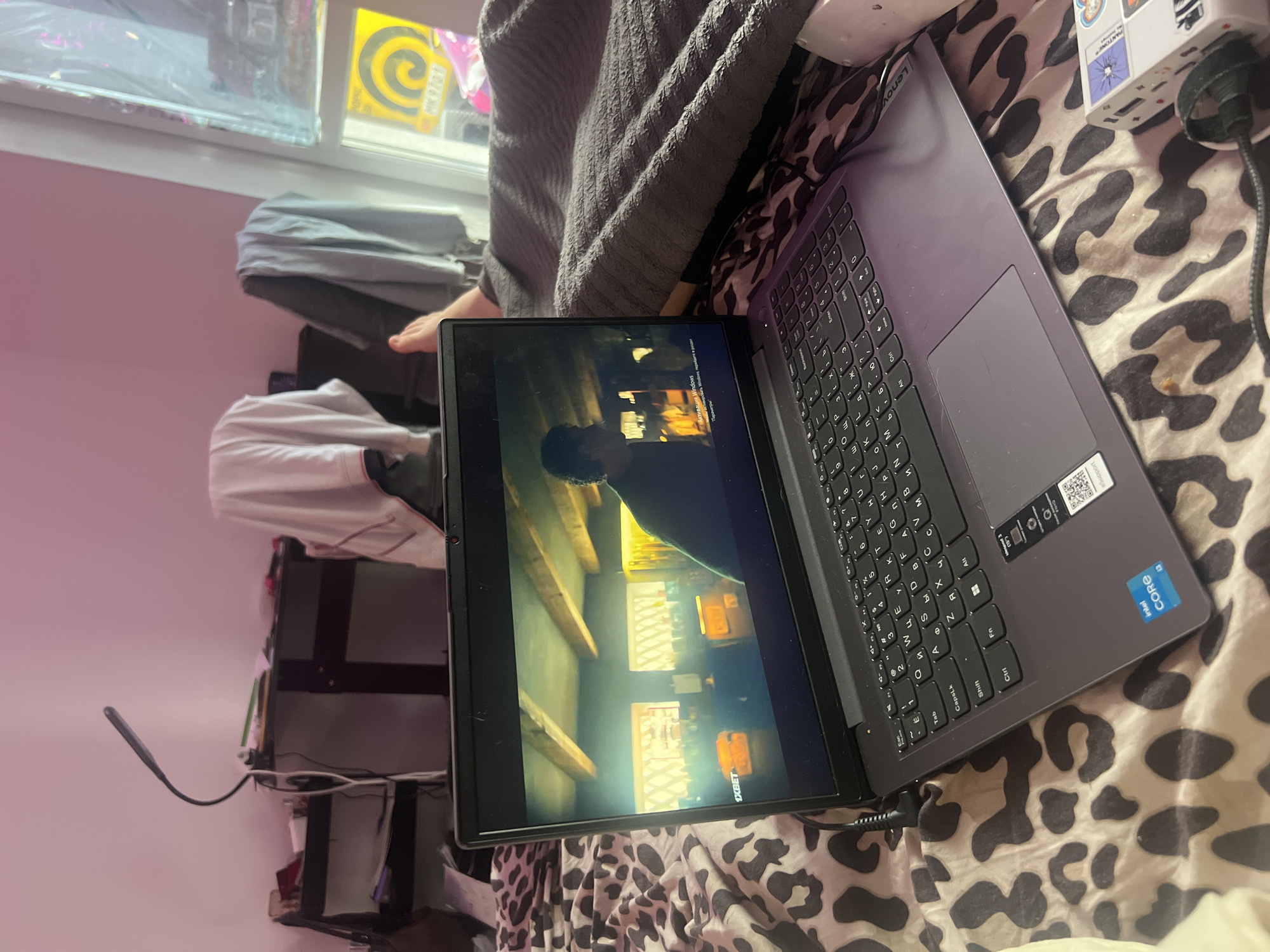 A Lenovo laptop is on a bed covered in a black and white animal print blanket. The laptop is open, and the screen is displaying a scene that includes what appears to be a vaulted ceiling with exposed beams and a person standing beneath them. The laptop is plugged in with a power adapter. To the left of the laptop, a small gooseneck lamp is plugged into a shelf unit. The room has a pink wall and a window in the upper left corner. The window has a yellow object with swirling markings, and clothes are draped over the shelves. In the upper right corner, a power strip is visible with stickers on it. A person is sitting on the bed behind the laptop.