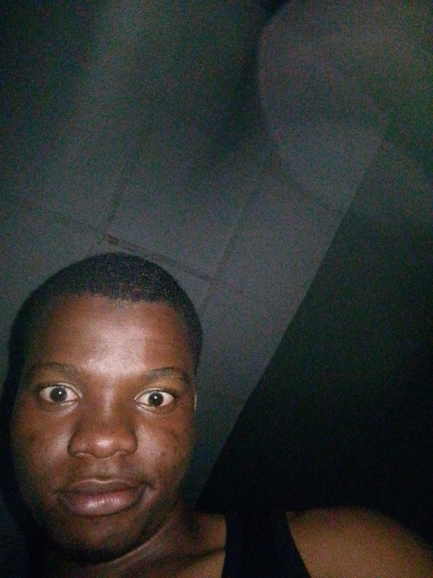The image is a close-up, selfie-style photograph of a Black male subject. The subject's face occupies the majority of the frame, with his eyes wide open and looking directly at the camera. His mouth is slightly open, with his upper lip appearing relaxed and his lower lip slightly pursed. The lighting is dim, creating a grainy texture throughout the image and casting shadows that obscure details in the background. The subject appears to be indoors, as suggested by the textured, dark background that resembles a wall or fabric. There are no discernible objects or other individuals present in the frame. The image does not provide any specific visual cues to determine the exact location within Ipetumodu, Nigeria.