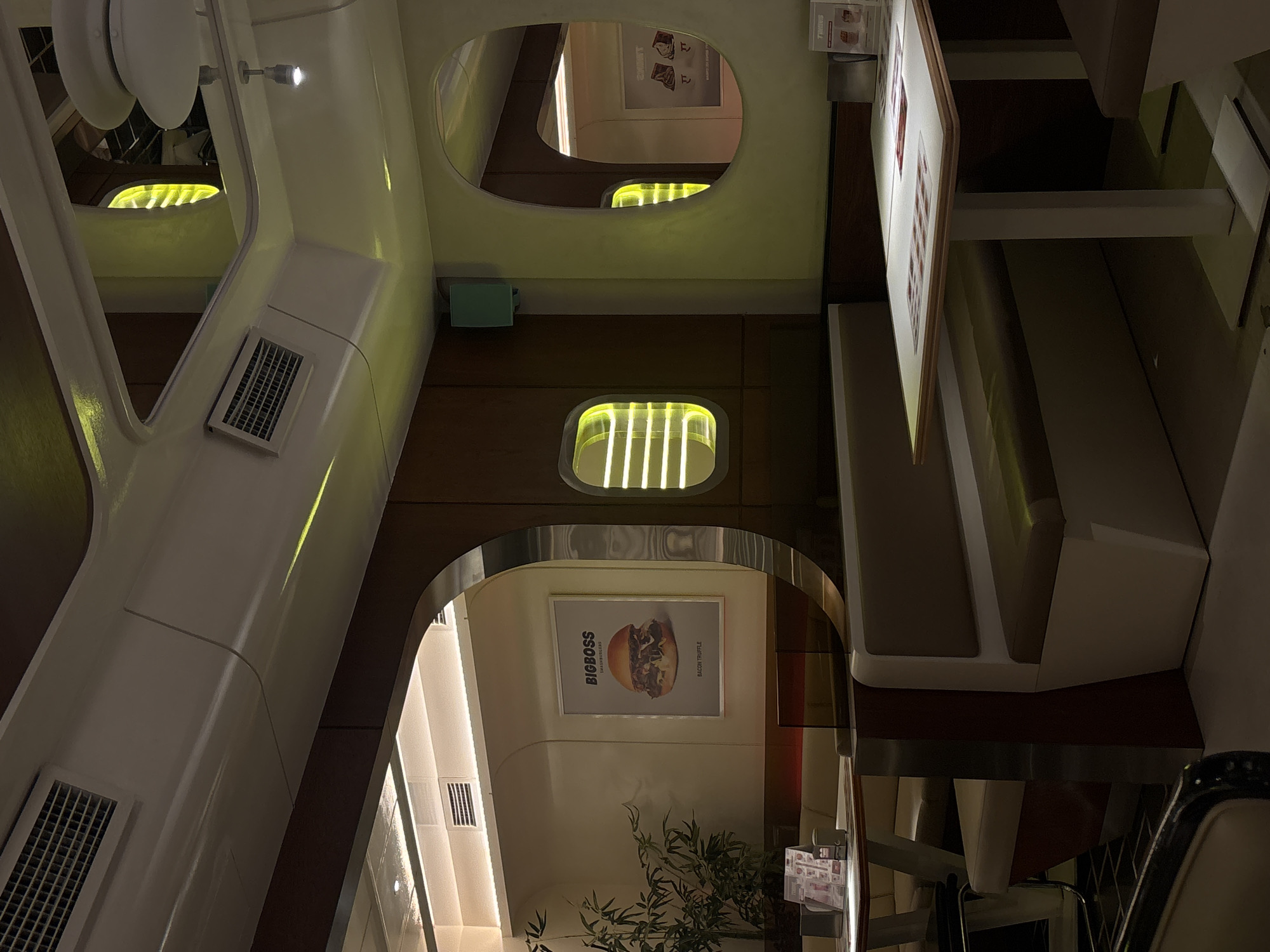 This image captures the interior of a contemporary restaurant or diner in Yerevan, Armenia, characterized by its sleek, modern design with a potential retro-futuristic or transportation-inspired aesthetic. The space is currently empty of people, suggesting a quiet moment.
The architecture features predominantly smooth, curved white walls and ceilings that create a sense of enclosed fluidity. These are contrasted with sections of dark, polished wooden paneling, particularly along the lower walls and supporting structures. The lighting is artificial, casting a warm, yellowish-green glow throughout the space. This light emanates from recessed linear strips integrated into the ceiling and walls, as well as from distinctive rectangular and oval-shaped back-lit panels that resemble windows. Additional white spotlights provide task lighting.
Visible objects include multiple rectangular ventilation grilles integrated into the white wall sections. Along the right side of the frame, comfortable booth seating with light-colored (beige or grey) upholstery is arranged, accompanied by dark wooden table surfaces.
A prominent decorative element is a large framed poster on a far wall, showcasing a delicious-looking burger. Visible text on this poster reads "BIGBOSS," "SMASHBURGERS," and "BACON TRUFFLE." Below the poster, a small, leafy plant with bamboo-like stalks adds a touch of greenery. In the upper right corner, an illuminated menu board is partially visible, though its specific contents are unreadable from this distance. Several oval reflective surfaces, possibly mirrors or polished metal, are embedded in the upper wall sections, offering distorted reflections of the interior and enhancing the sense of depth. The overall ambiance is clean, stylish, and inviting, with a strong emphasis on architectural design and integrated lighting.