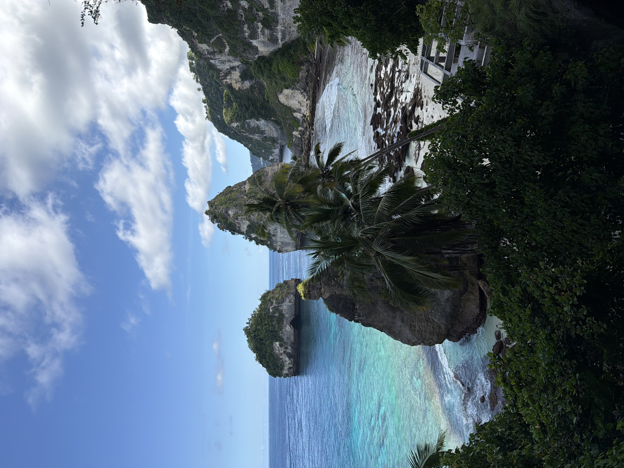 The image showcases a tropical coastal scene, likely in Pejukutan, Indonesia, given the limestone cliffs and turquoise waters. Two prominent rock formations, one with palm trees, are visible in the foreground. The water transitions from a deep blue further out to a vibrant turquoise closer to the shore. A rocky beach is present in the distance. Dense, green vegetation frames the right side of the image, while the sky is filled with white, fluffy clouds against a blue backdrop. Part of a man-made structure is visible above the rocky beach.