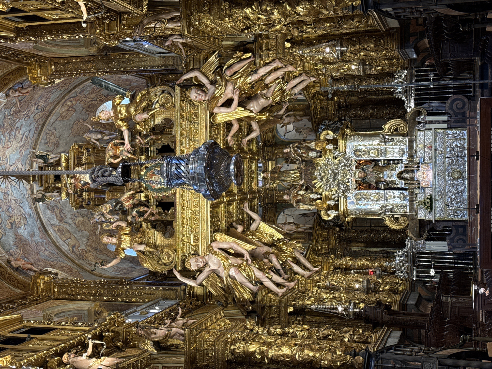 This is an interior view of a richly decorated church, likely a cathedral, in Santiago de Compostela, Spain. The scene is dominated by an ornate altar and retablo, extensively gilded and adorned with numerous sculptures. In the foreground, dark wooden pews are visible, suggesting the nave of the church.
The retablo is a monumental structure featuring a central niche containing a statue, likely of a saint, surrounded by a profusion of sculpted figures and intricate gold leaf ornamentation. Above the central area, several larger gilded sculptures of winged figures, resembling angels or cherubs, are suspended or mounted, creating a dynamic and theatrical display. Further up, more sculptural elements and painted frescoes adorn the vaulted ceiling. A large, ornate silver lantern hangs from the ceiling in the center of the frame, adding to the grandeur.
The overall impression is one of overwhelming opulence and religious devotion, characterized by the extensive use of gold, elaborate carvings, and a multitude of religious iconography. The lighting appears to be a combination of ambient church light and possibly some artificial lighting illuminating the altar and surrounding decorations.