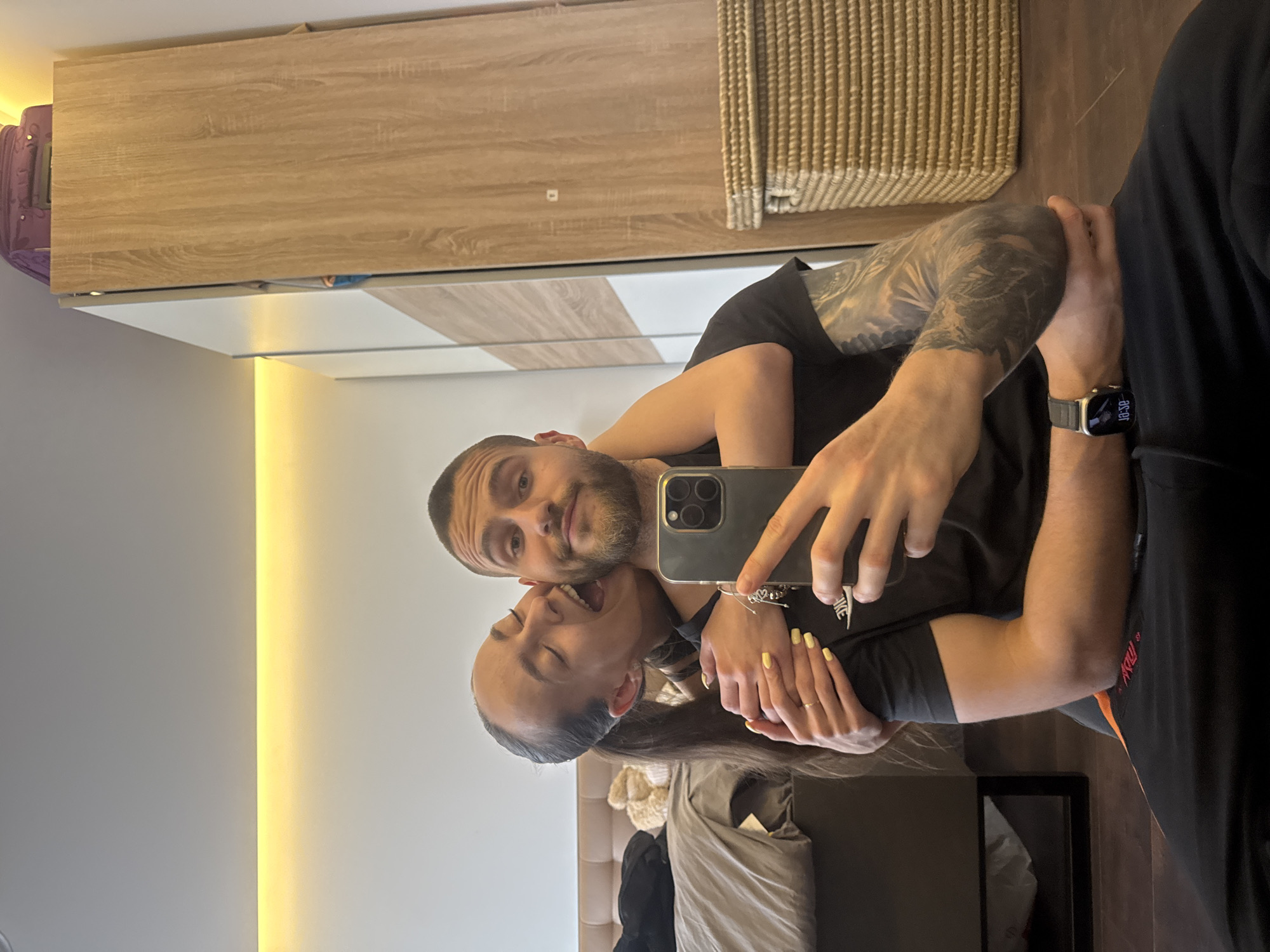 A man and a woman are posing for a selfie in front of a mirror. The man, on the right, is wearing a black shirt and a smart watch. He has a beard and short hair. The woman, on the left, has long dark hair and is wearing a black shirt. The location appears to be indoors, possibly a bedroom, with a bed and furniture visible in the background. There are no specific landmarks or objects that would identify the location as Sofia, Bulgaria.