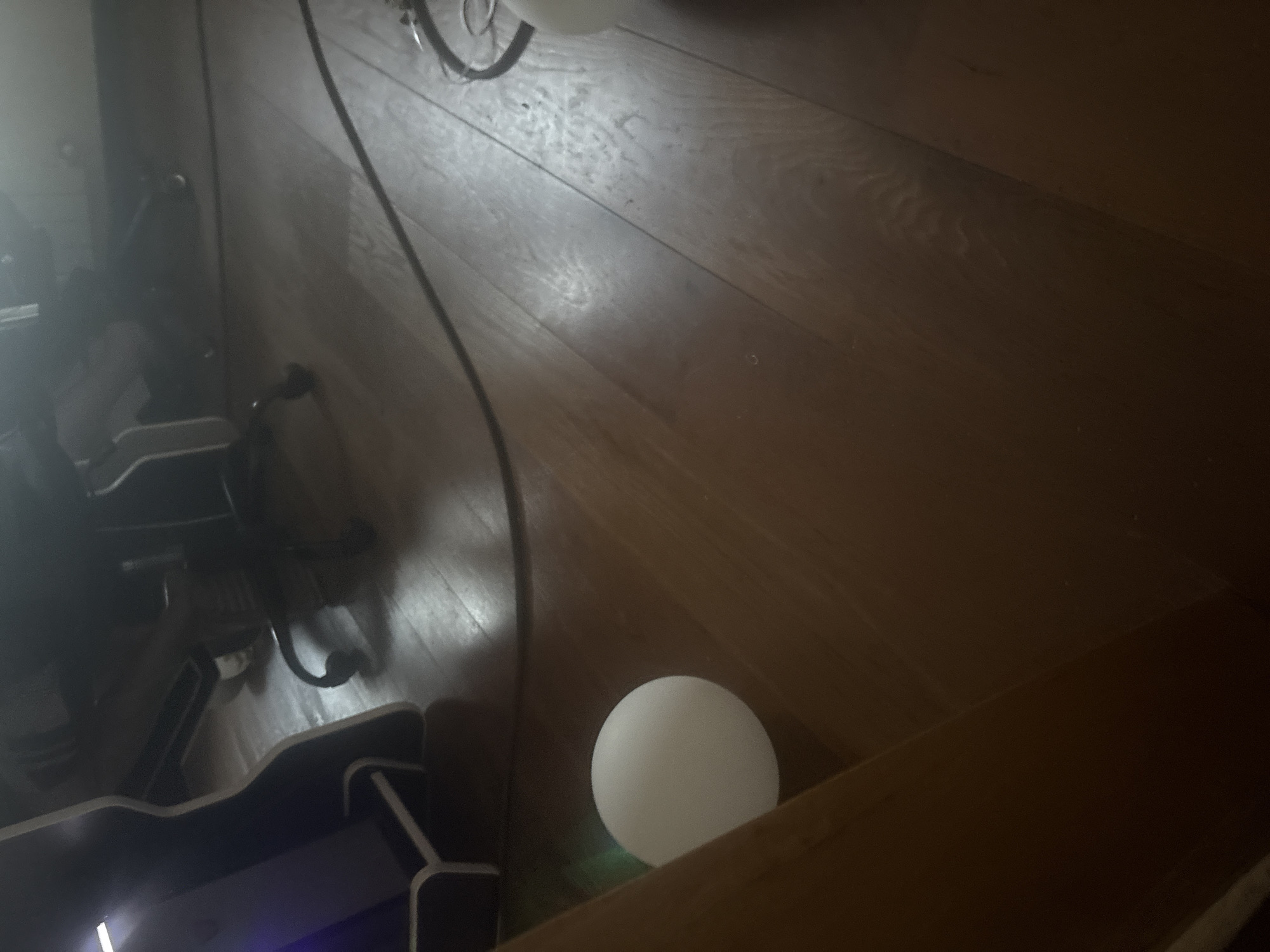 The image shows a dimly lit interior space with a wooden floor. In the foreground, a white spherical object, possibly a ball, is visible on the left side. To the right, a black electrical cord snakes across the floor. In the background, the base of a desk or computer tower is visible on the far left, and parts of a dark office chair with wheels can be seen. The scene appears to be a domestic or office environment. No specific details about the city of Orekhovka, Russia, can be inferred from the image itself.