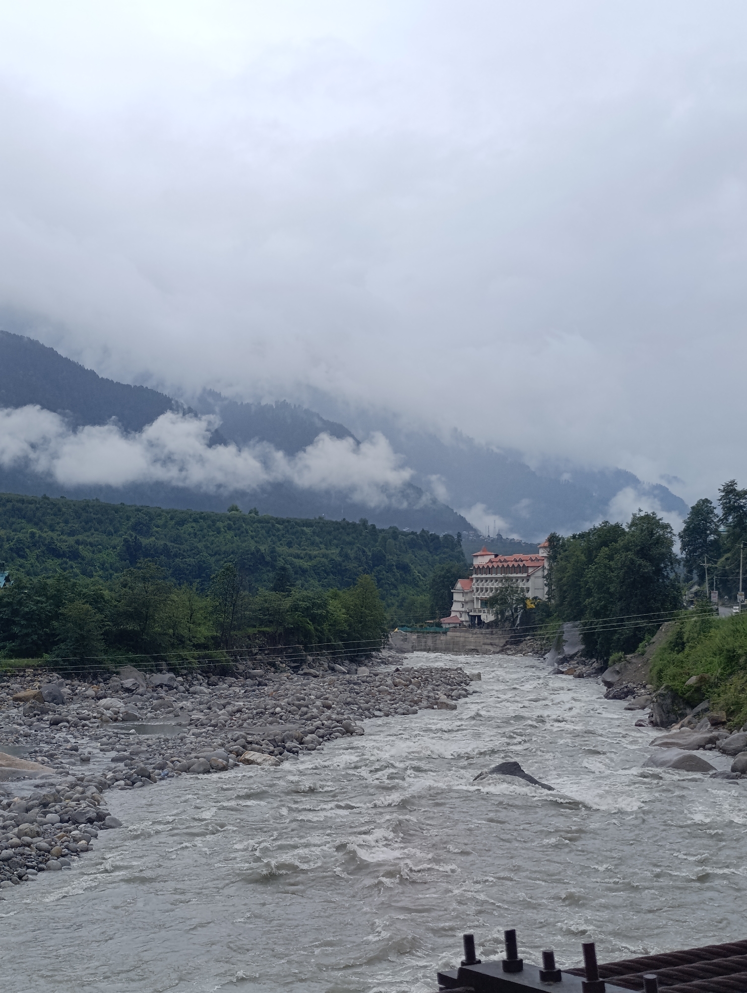 The image depicts a wide, turbulent river flowing through a mountainous, forested landscape in Kalath, India. The river, with its grey, choppy water, occupies the foreground and middle ground, with a rocky, grey shore visible on the left bank. A large, multi-story building with a distinctive red-tiled roof and white facade, resembling a hotel, is situated on the right bank, nestled amongst lush green trees. Behind the building, steep, tree-covered hills rise, partially obscured by thick, white clouds that cling to the mountain slopes. The sky is overcast and grey, contributing to the moody atmosphere of the scene. The far background shows more mountains fading into the mist. The scene is natural and serene, with no people or animals visible.