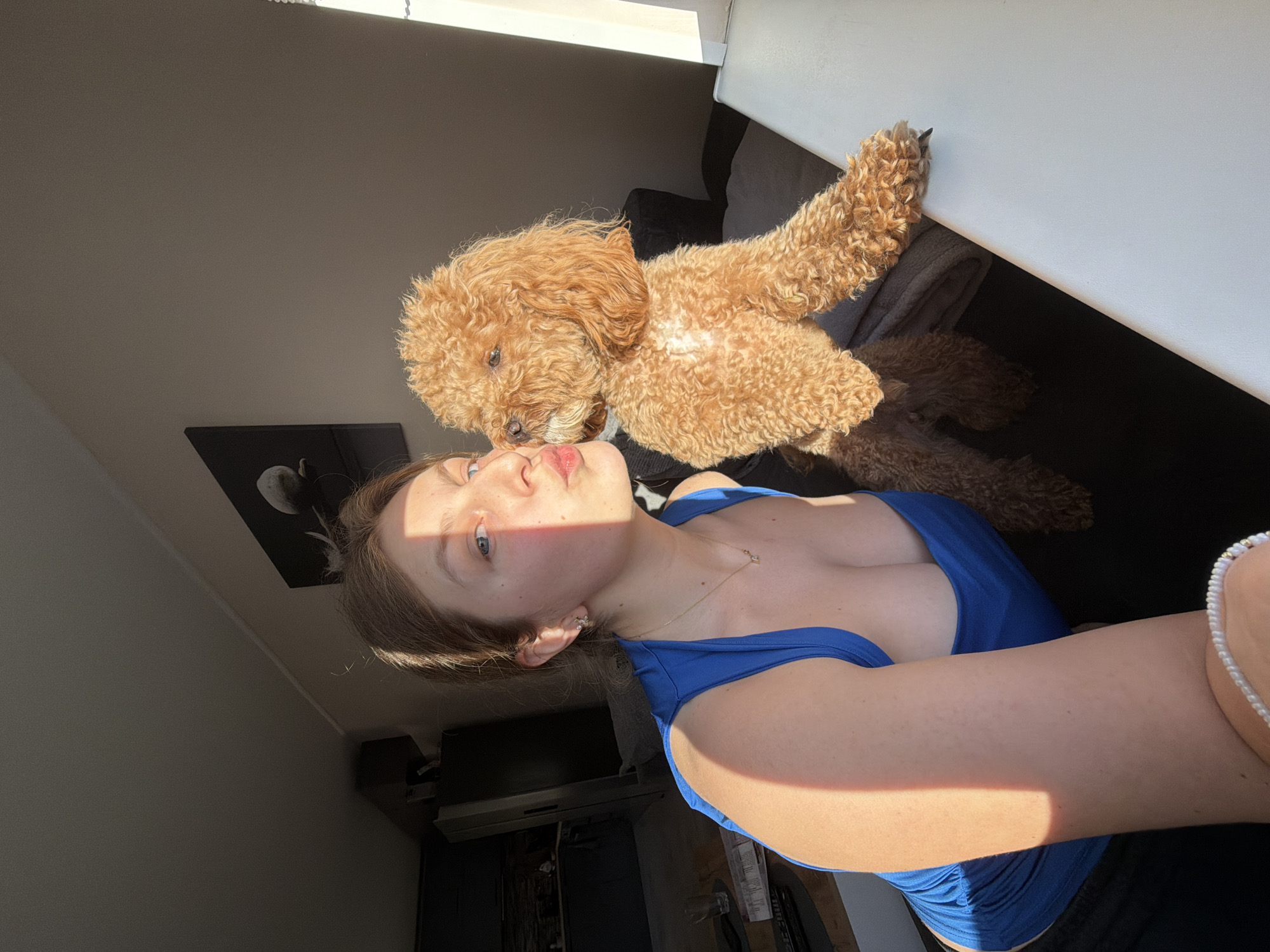 A young woman with light skin, light brown hair, and blue eyes is taking a selfie with a light brown poodle. The woman is wearing a blue top. The dog is positioned above the woman's face and appears to be resting on a surface. The photo is taken indoors with a window in the background, providing natural light. There is artwork visible on the wall. The location is unknown and there is nothing in the image that associates to the city of Litakovo, Bulgaria.