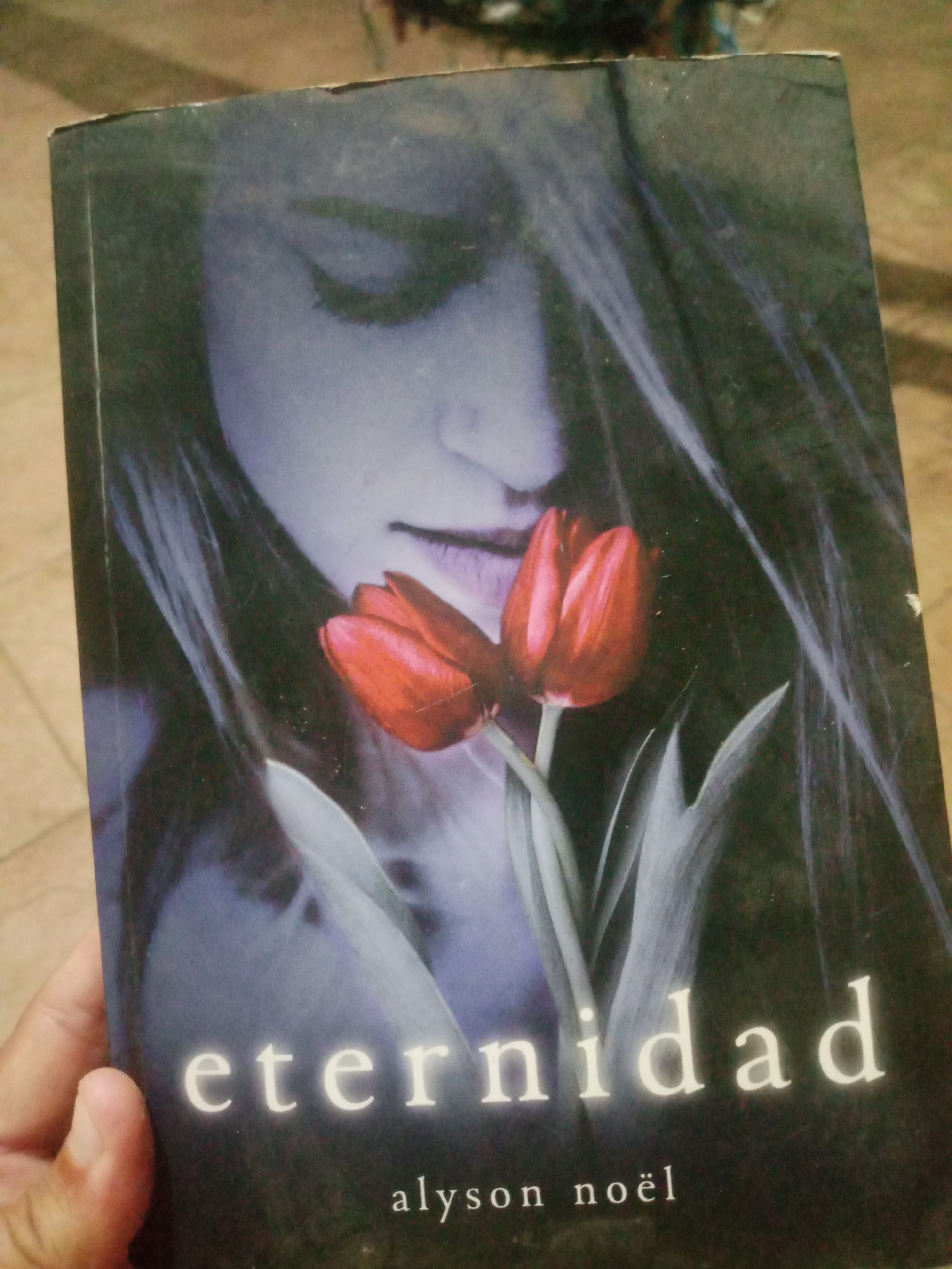 The image shows a hand holding a book with a dark cover. The book features a close-up of a woman's face, with her eyes closed and hair flowing. Two red tulips are positioned in the foreground, partially obscuring her face. The title of the book, "eternidad," is prominently displayed in white lettering at the bottom of the cover, followed by the author's name, "alyson noël." The background of the image is blurred and appears to be an indoor setting with a tiled floor. The lighting suggests it is daytime. There is no visible indication of the specific location within Santa Ana, Venezuela.