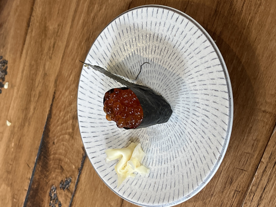 The image presents a single piece of Ikura Gunkan Maki (salmon roe sushi) and a small portion of pickled ginger, centrally positioned on a round, light-colored ceramic plate. The plate features a patterned surface with dark grey radial lines originating from its center. The sushi consists of a dark green nori (seaweed) wrap enclosing a base of rice, topped with a generous quantity of translucent, glossy, orange-red salmon roe. A small amount of pale yellow, thinly sliced pickled ginger (gari) is situated to the left of the sushi on the plate. The plate rests directly on a smooth, warm-toned wooden surface, exhibiting distinct grain patterns. A few minute, light-colored specks are visible on the wooden background. No individuals or specific architectural features are present to identify a precise location within Mexico City, Mexico.