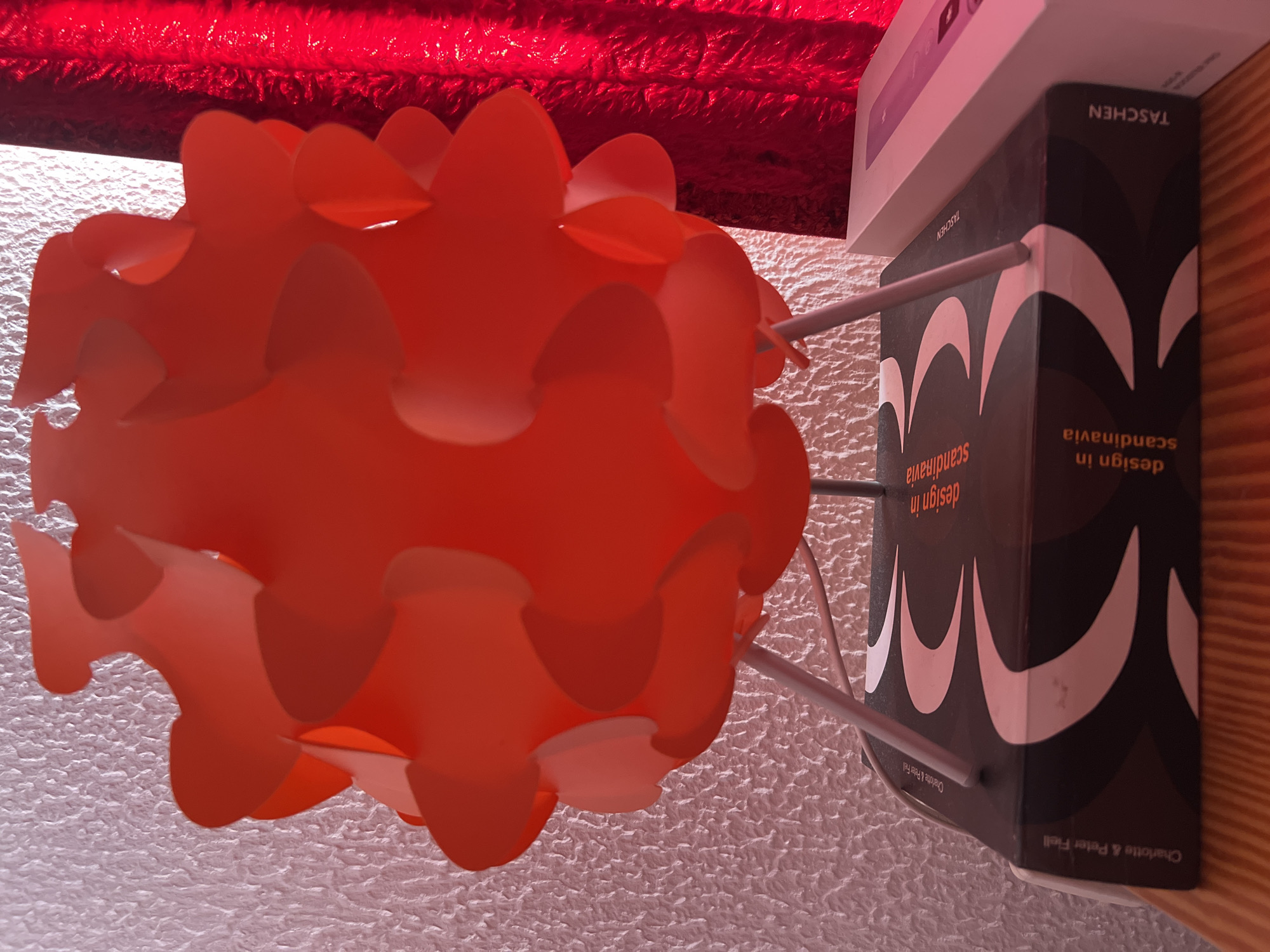 A red, spherical, multi-paneled lamp sits on top of a stack of books. The lamp has a slightly textured surface and is lit from within. The books are stacked horizontally, with the top book being dark grey and featuring a white wavy pattern on its cover. The title "design in scandinavia" is visible on the spine of the book. The words "Charlotte & Peter Fiell" are also present on the spine of the book on the left. The stack of books is on a wooden surface. To the right of the books, a white box with a pinkish hue is partially visible. The background consists of a textured white wall on the left and a red, crinkled fabric on the right, likely a curtain. The lighting in the scene is warm, casting shadows. The overall scene appears to be indoors, possibly on a shelf or table. This image was taken in Oroklini, Cyprus.