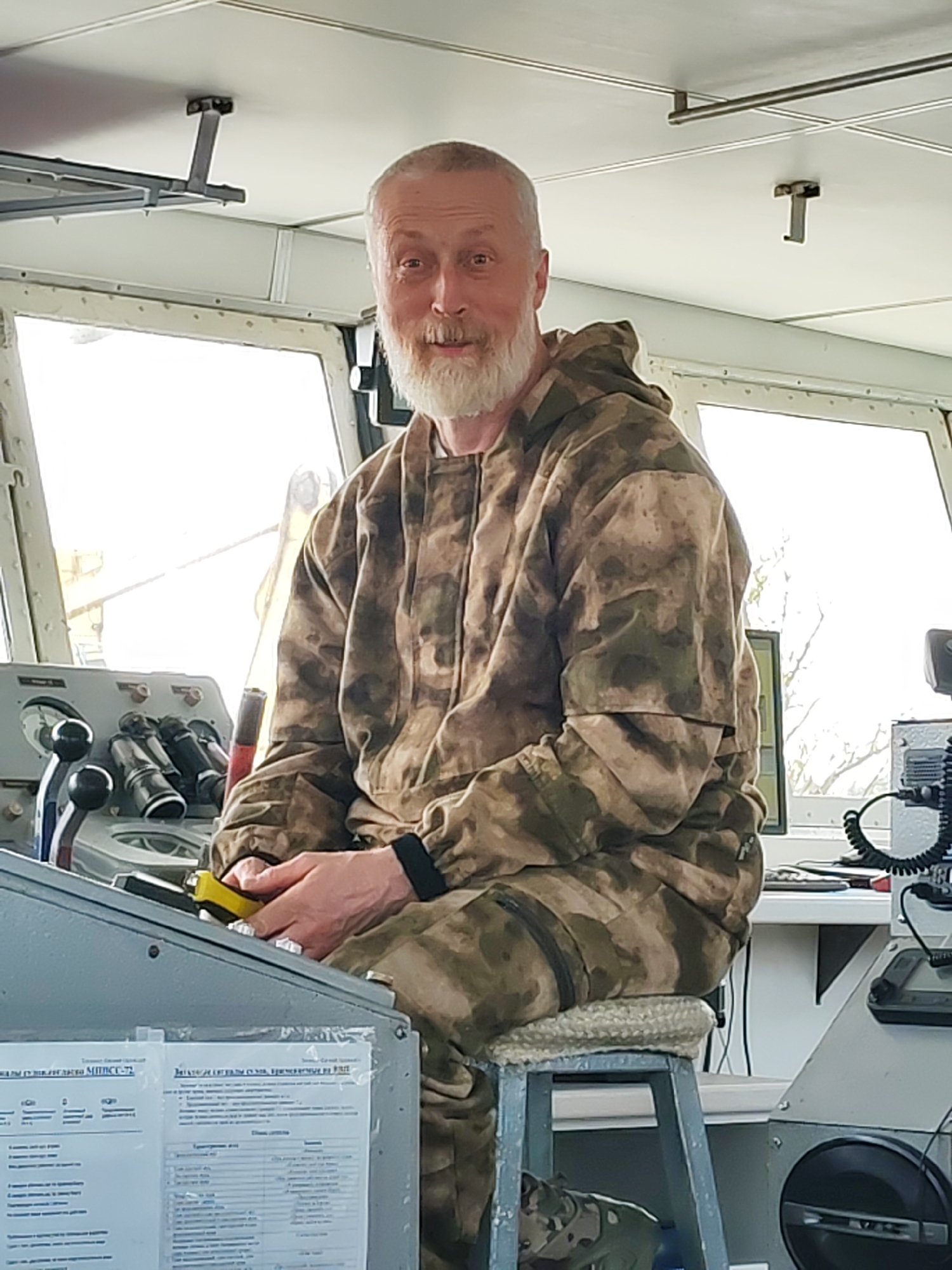 A middle-aged man with a white beard sits inside the bridge of a boat. He is wearing a camouflage hooded jacket and pants. The boat's bridge is filled with navigation equipment. Outside the window, there is a glimpse of a cityscape. The boat is located in Yakhroma, Russia.